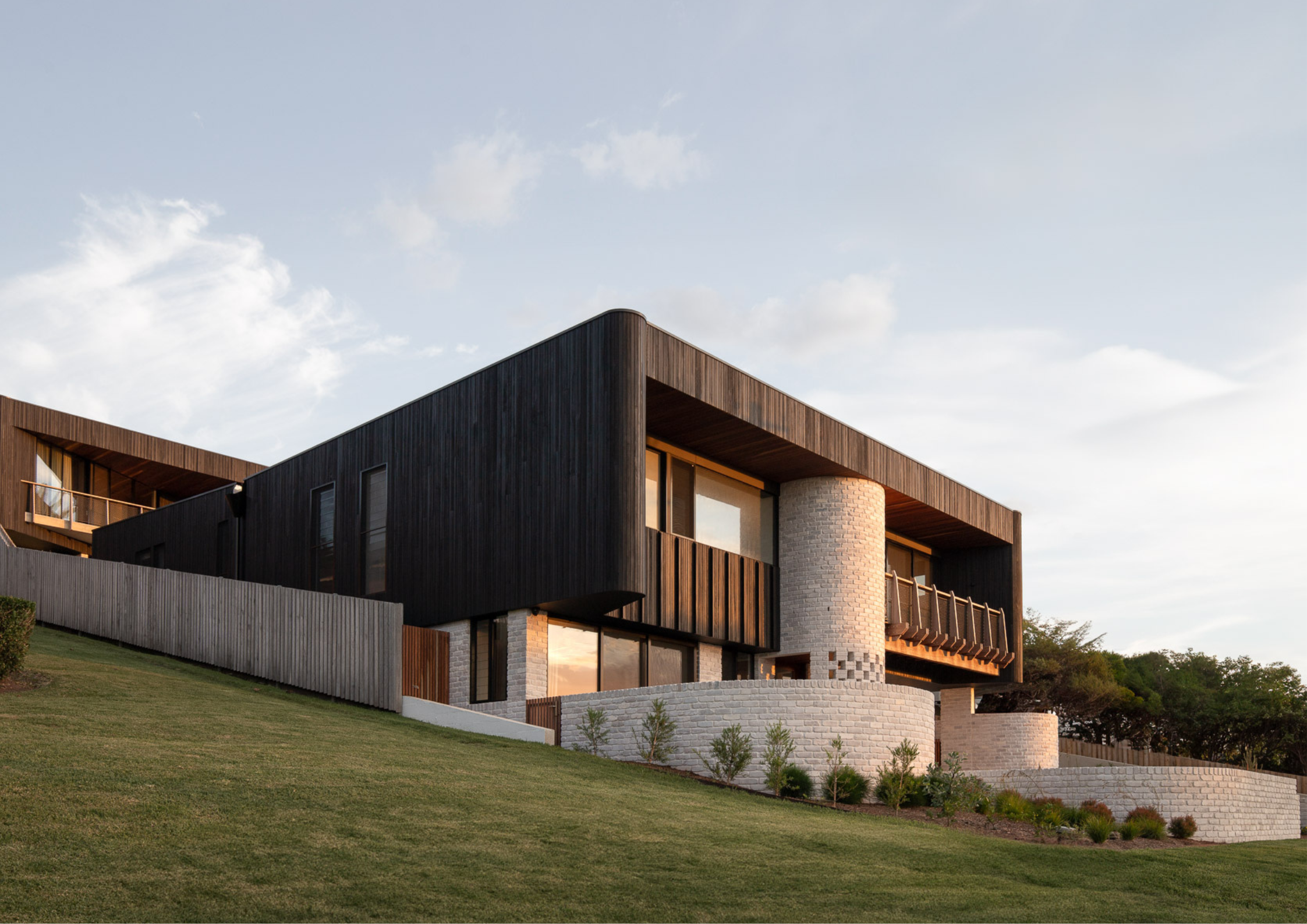 Modern hillside residence with dark timber cladding, curved brick detailing, and expansive balconies overlooking landscaped lawns.