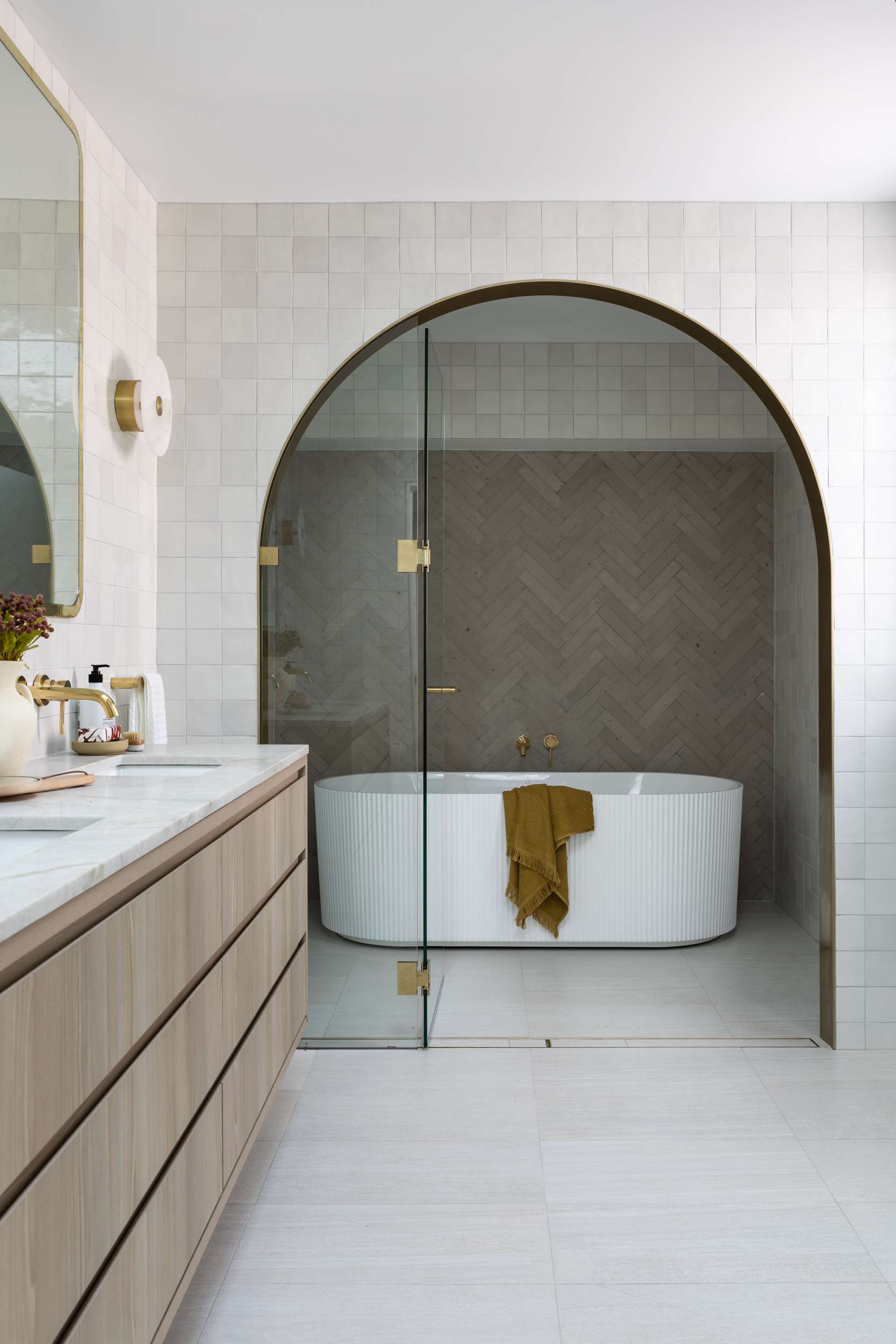 Minimal bathroom with arched shower enclosure, white tiled walls and ribbed freestanding bath with brass detailing.