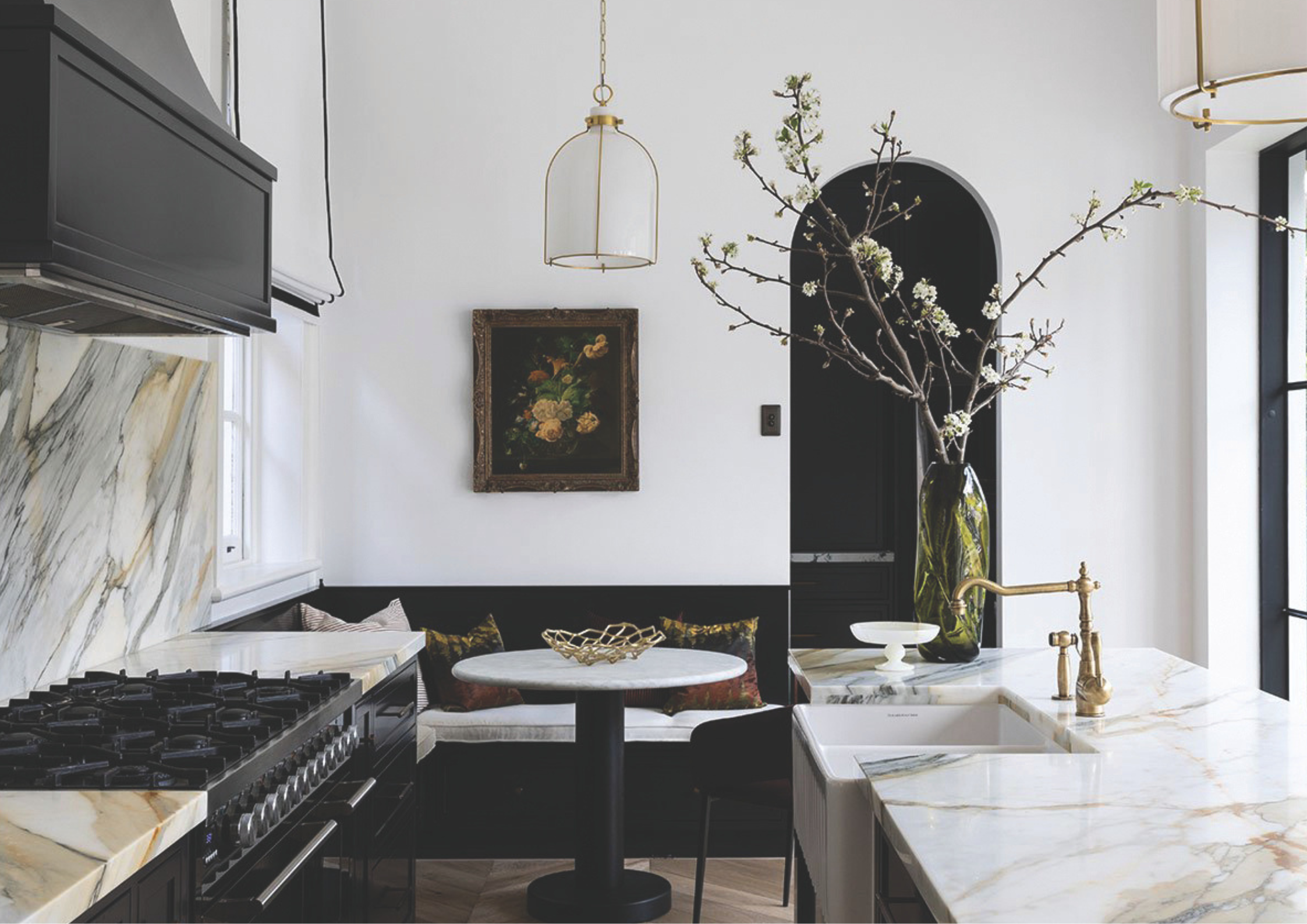 Elegant black and white kitchen with marble surfaces, brass tapware, arched doorway and a built-in dining nook styled with a flowering branch arrangement.