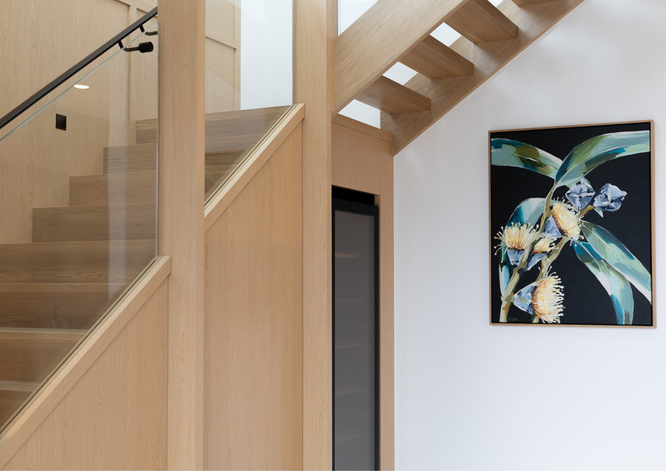 A light-filled interior with a timber staircase, glass balustrade, and a framed artwork on a white wall.