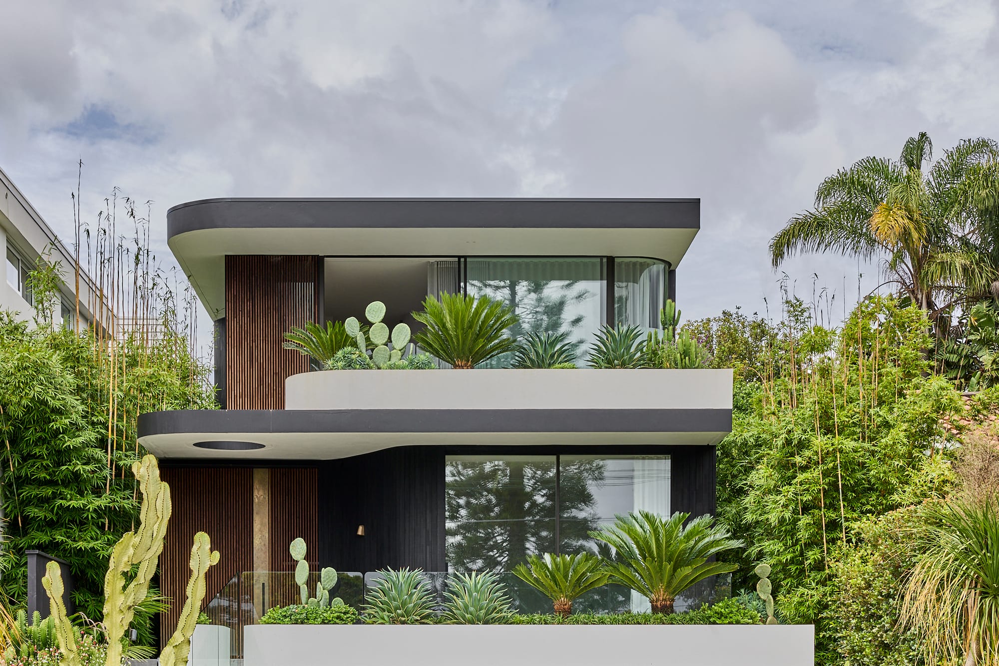 Contemporary two-storey home with curved concrete balconies, dark timber cladding, and lush cactus-filled planter beds surrounded by tropical landscaping.