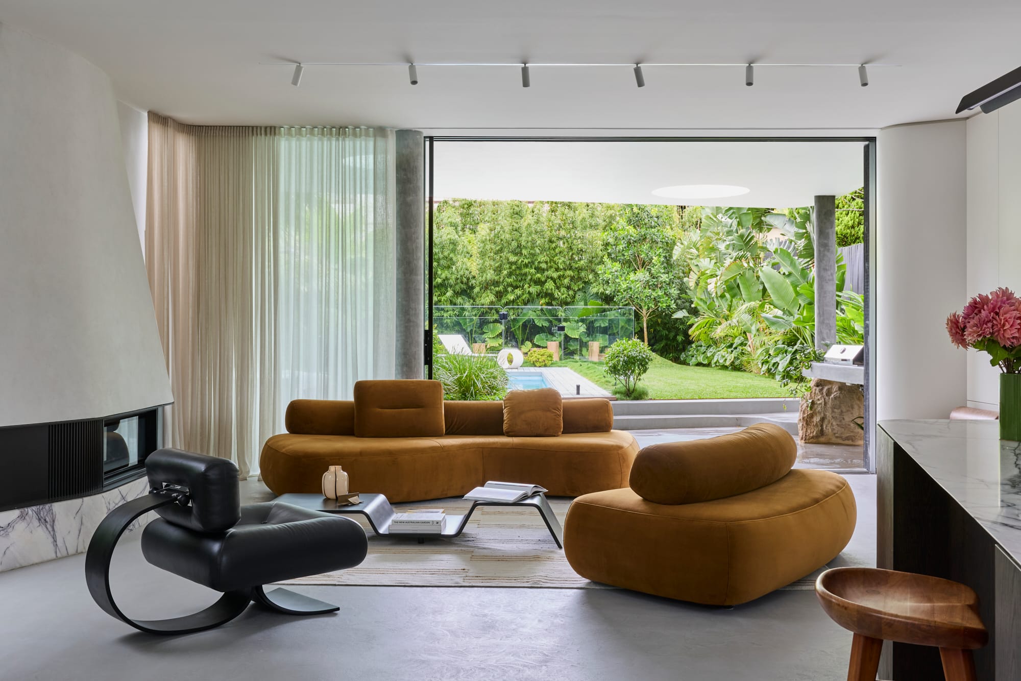 Minimal contemporary living room with sculptural curved sofas, soft natural light, and large openings connecting to a lush garden and pool.