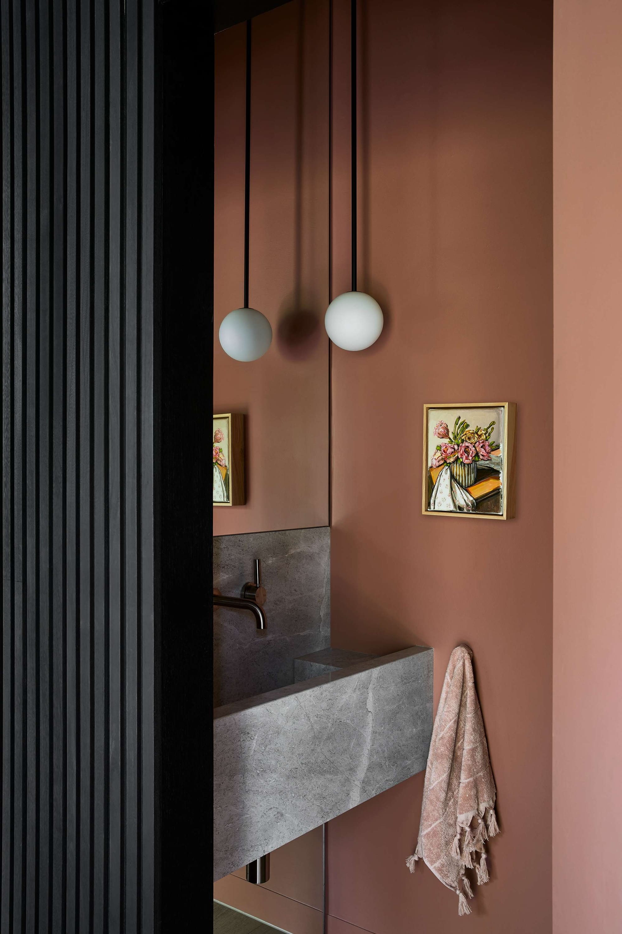 Contemporary powder room with blush-toned walls, a floating grey stone basin, spherical pendant lights, and framed floral artwork, partially screened by black vertical timber battens.