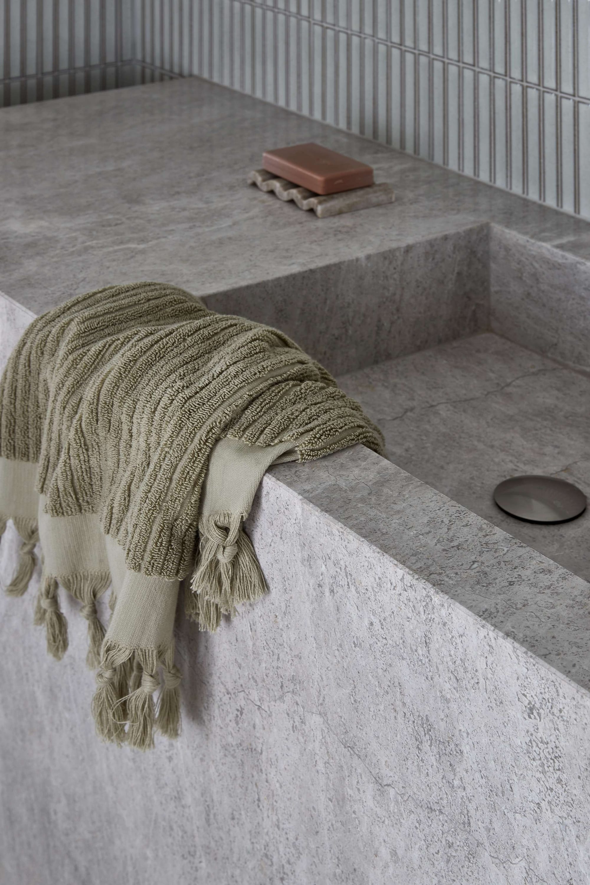 Minimalist stone bathroom detail featuring a monolithic grey stone basin, vertically stacked mosaic wall tiles, and a fringed olive-green towel draped over the edge, creating a calm, tactile composition.