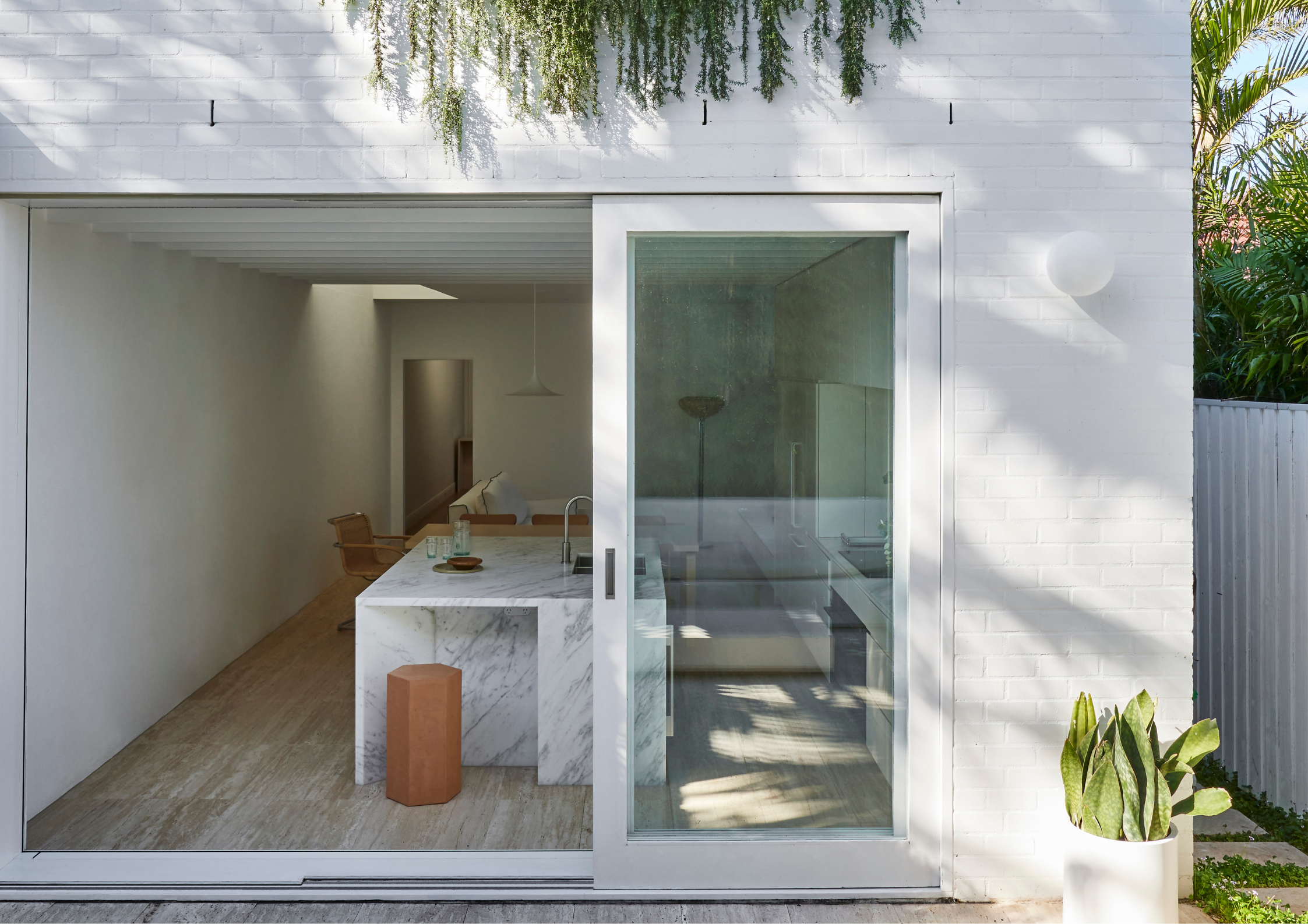 Minimal white brick extension with large sliding glass doors, a marble kitchen island, and soft natural light filtering through greenery.