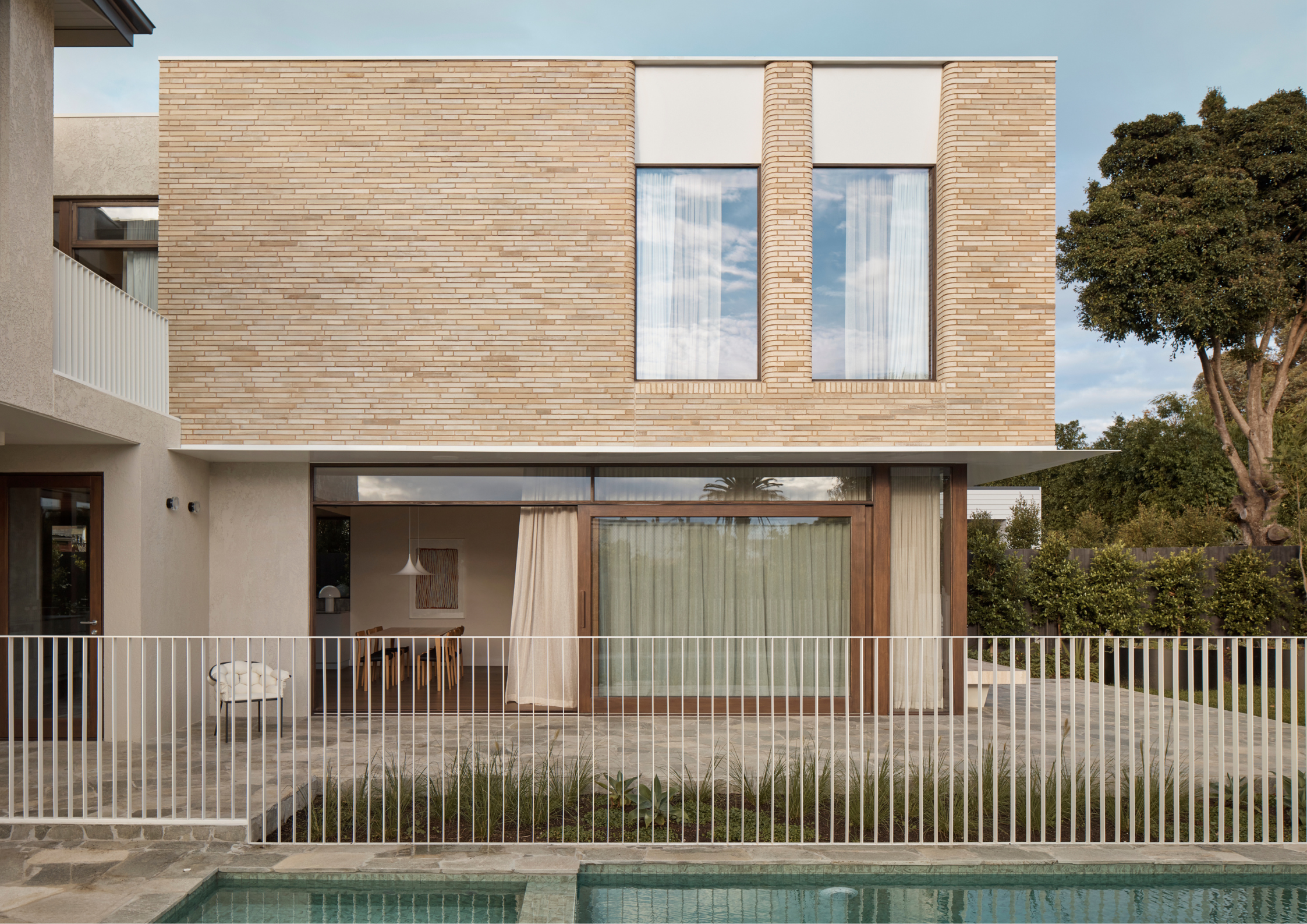 A contemporary home with a brick upper level, large windows, and a pool in the foreground behind a white fence.