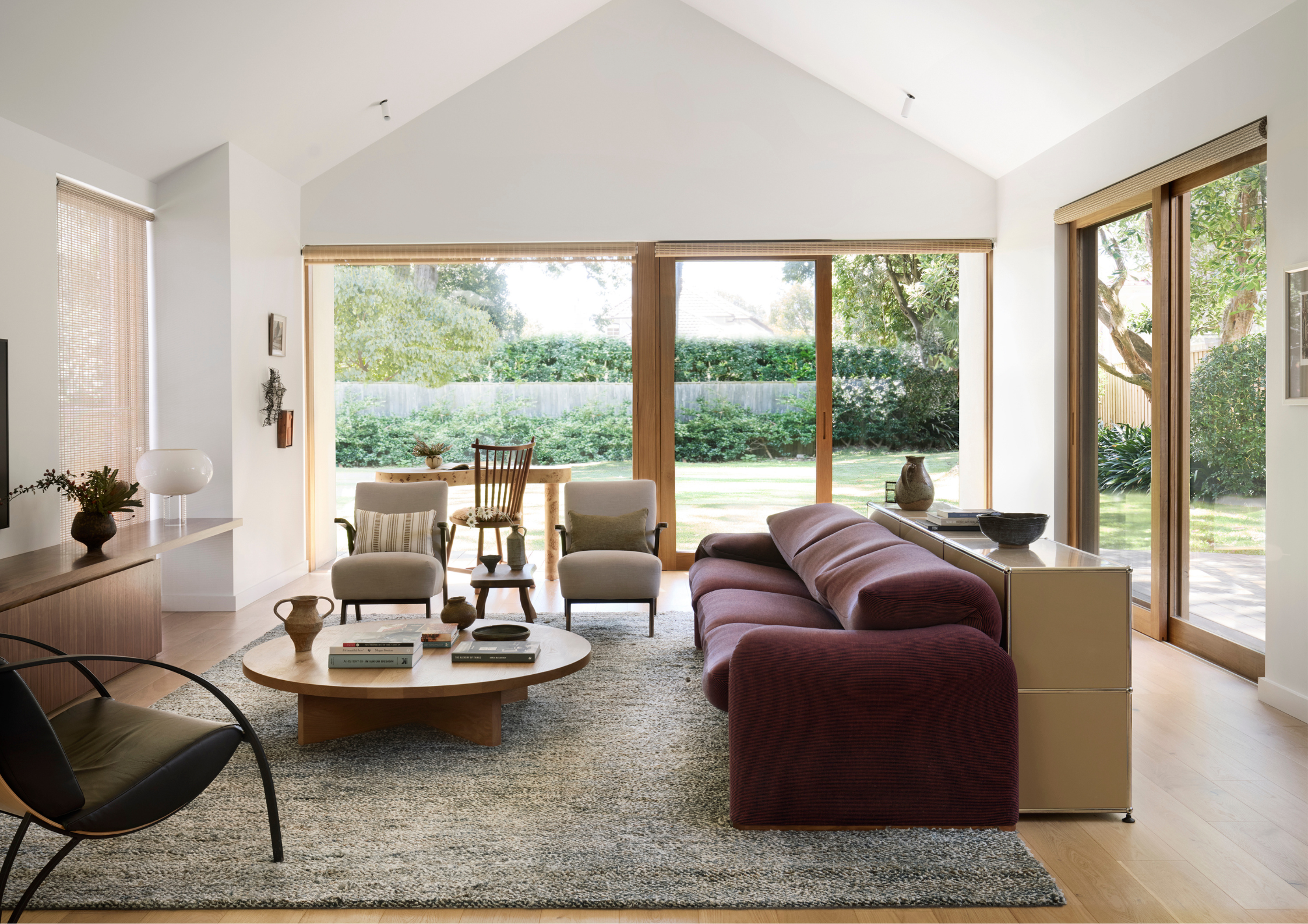 Bright contemporary living room with vaulted ceilings, large timber-framed windows, soft neutral furnishings and a curved burgundy sofa overlooking a lush garden.