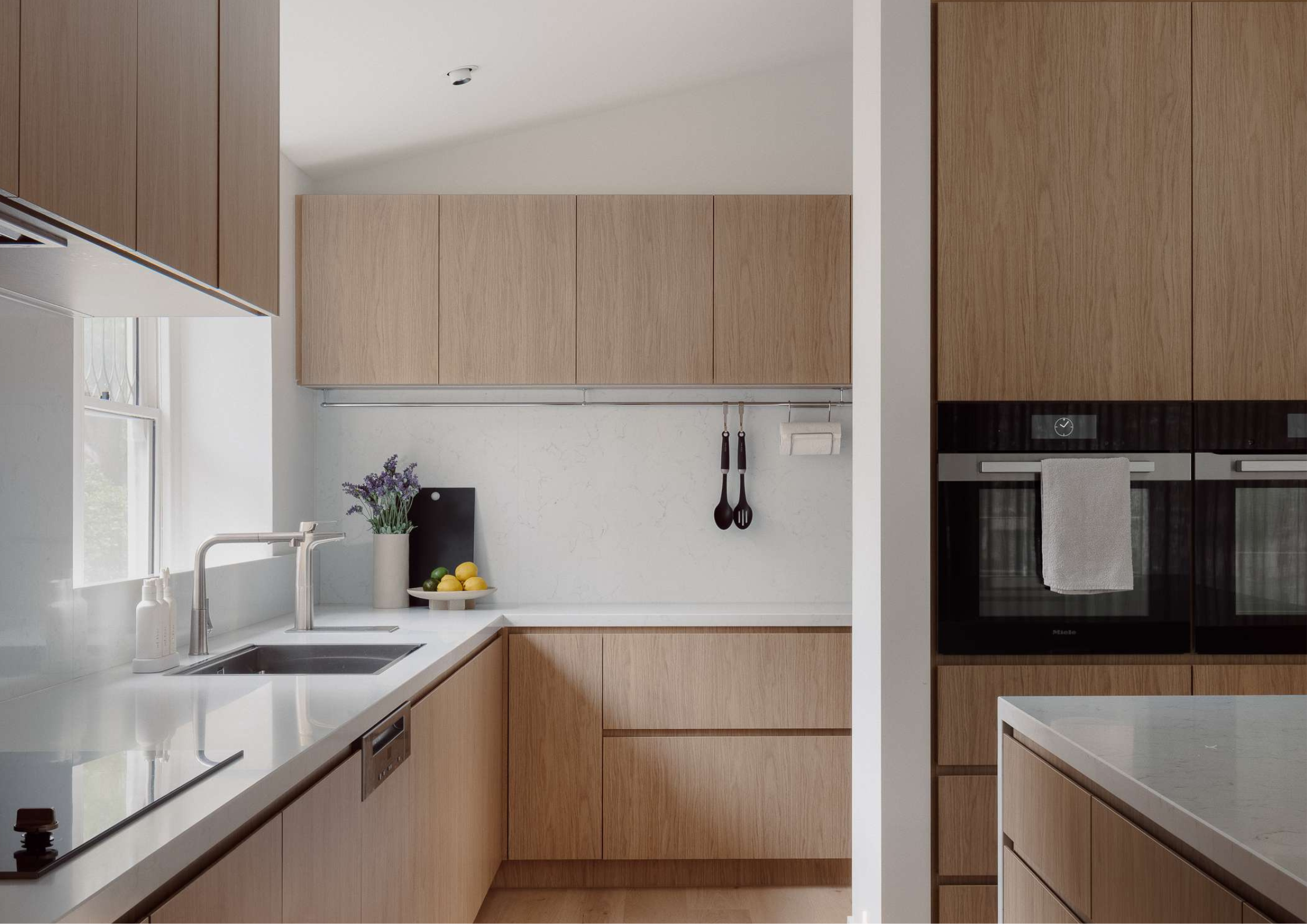 Minimal oak kitchen with concealed storage, white stone surfaces, and soft natural light.
