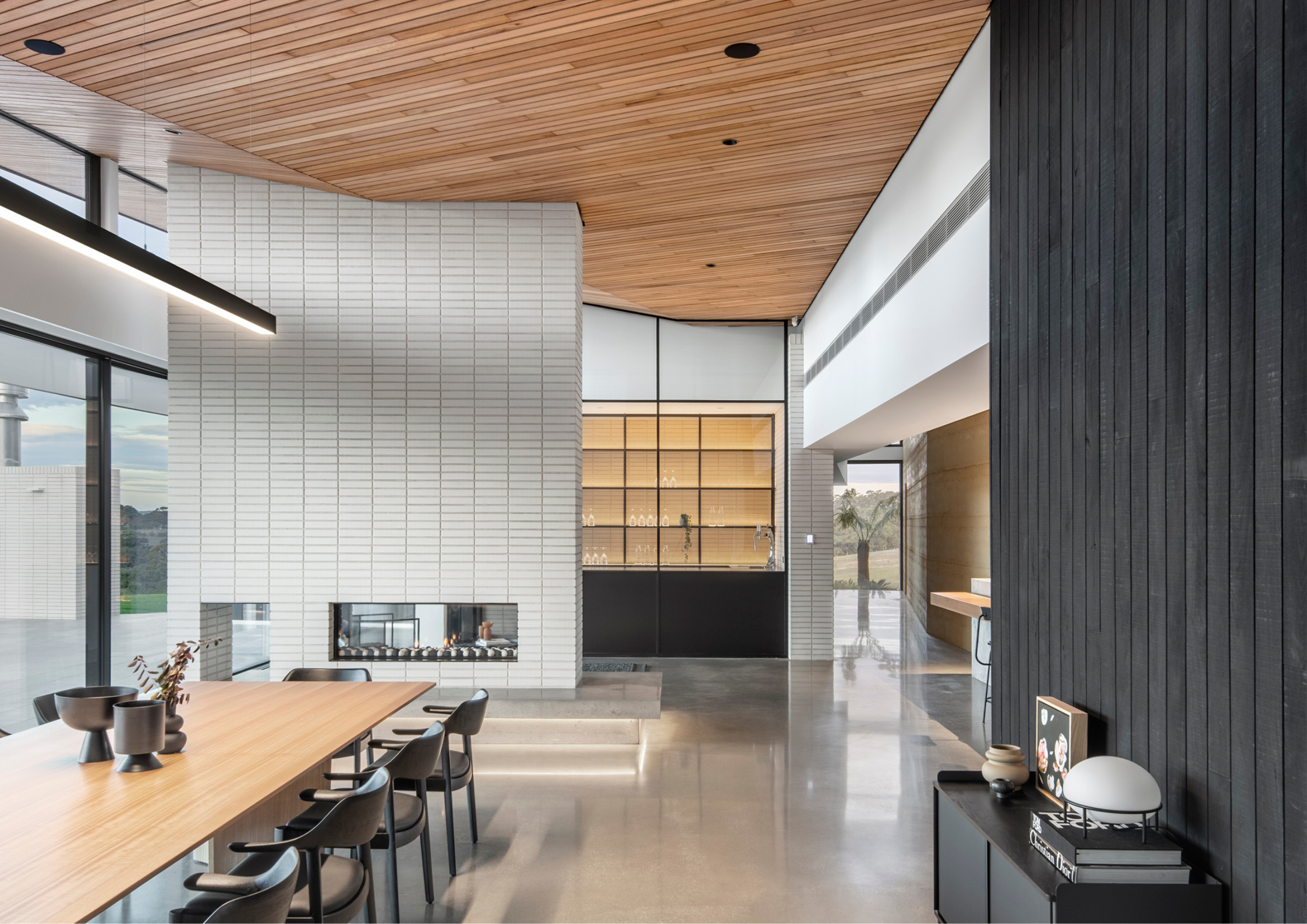 Contemporary open-plan dining space with timber-lined ceilings, white brick fireplace, polished concrete floors, and expansive glazing overlooking the landscape.