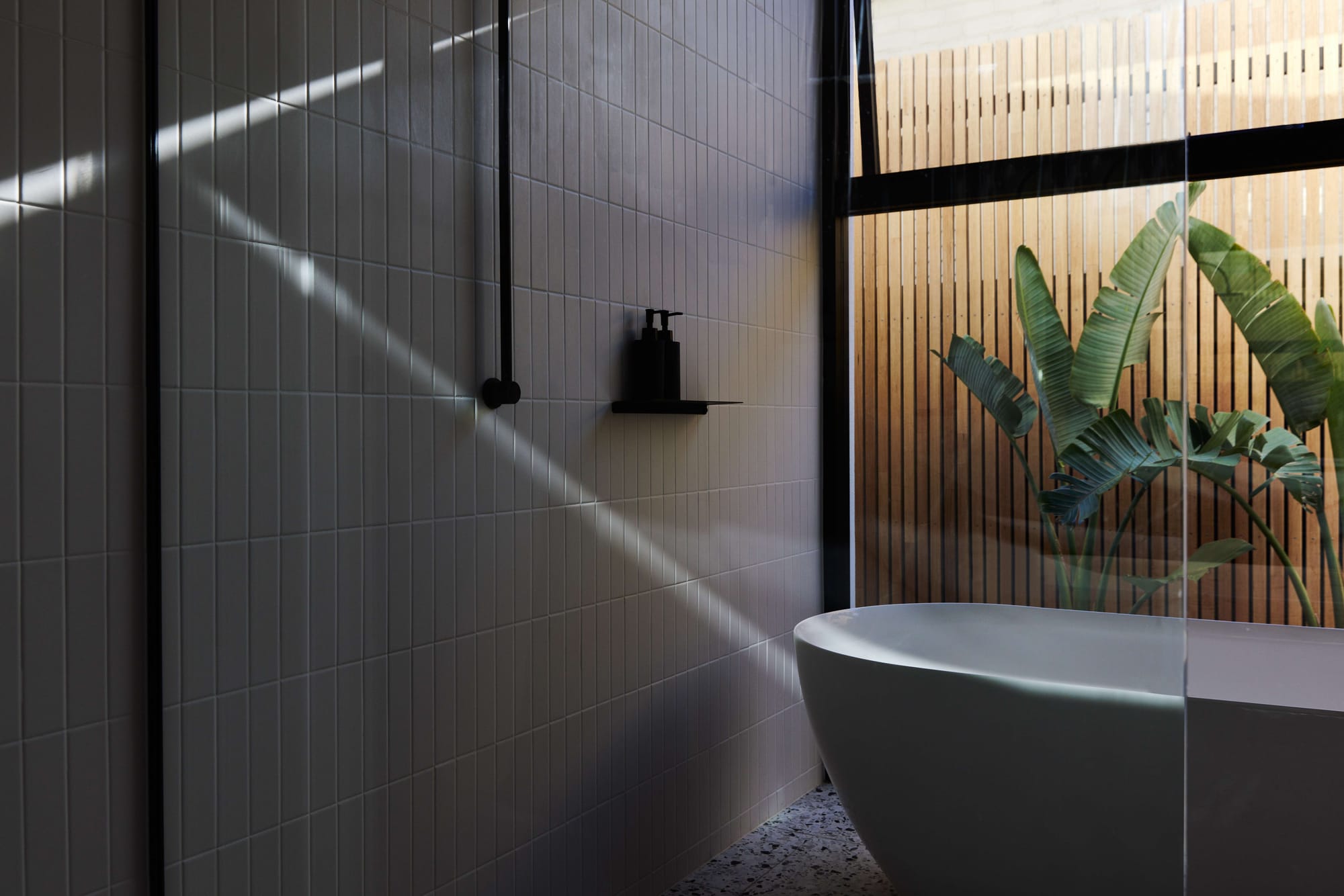 Minimal dark-toned bathroom with freestanding bath beside floor-to-ceiling glazing and tropical planting.