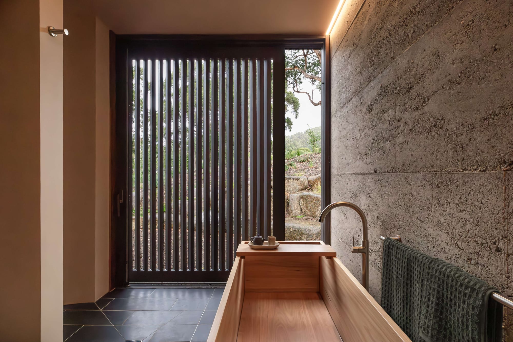 Japanese-inspired bathroom with timber soaking tub, rammed earth walls and slatted screen opening to native landscape views.