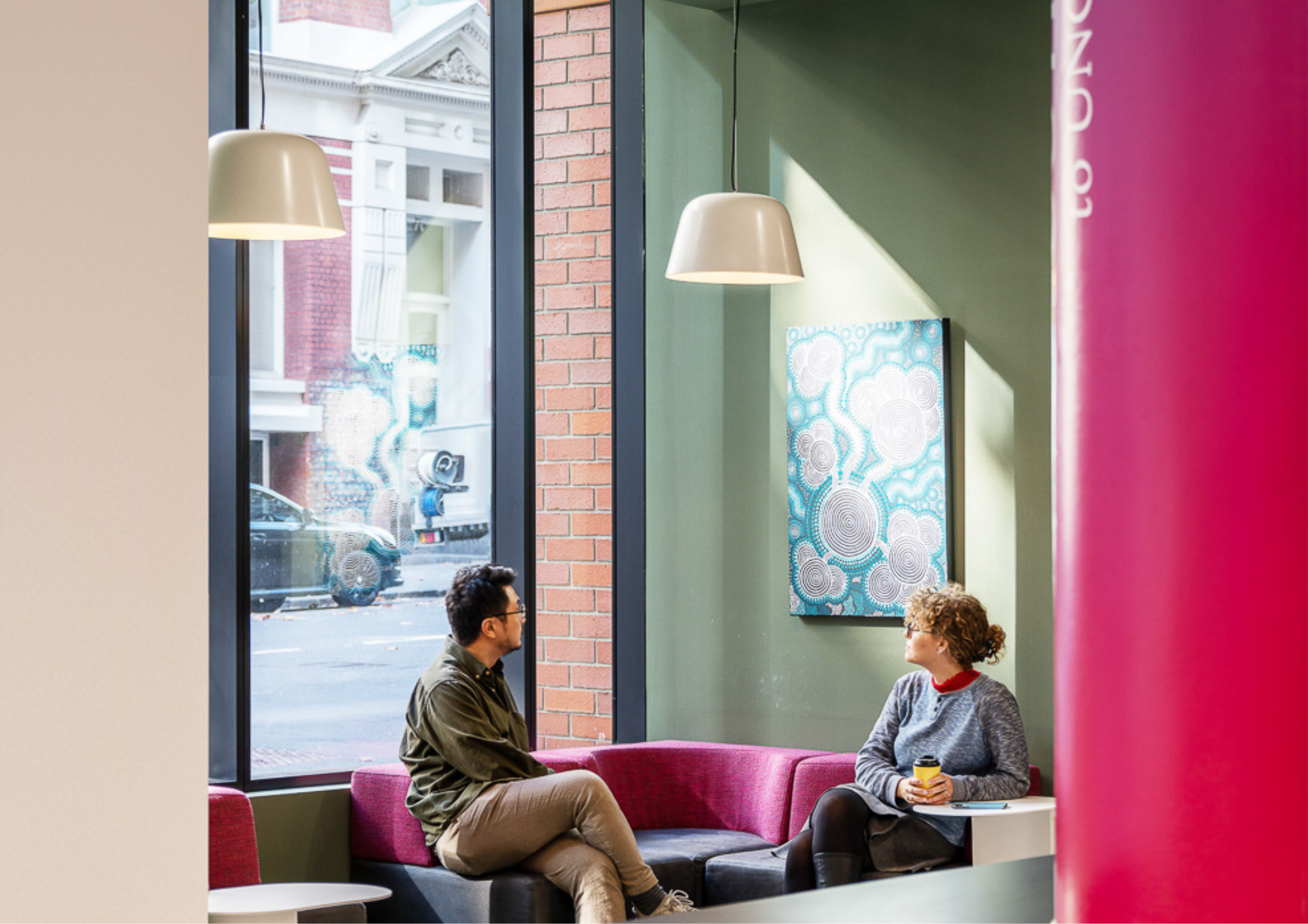Modern communal lounge with pink upholstered seating, tall street-facing windows, and soft pendant lighting creating a relaxed social atmosphere.