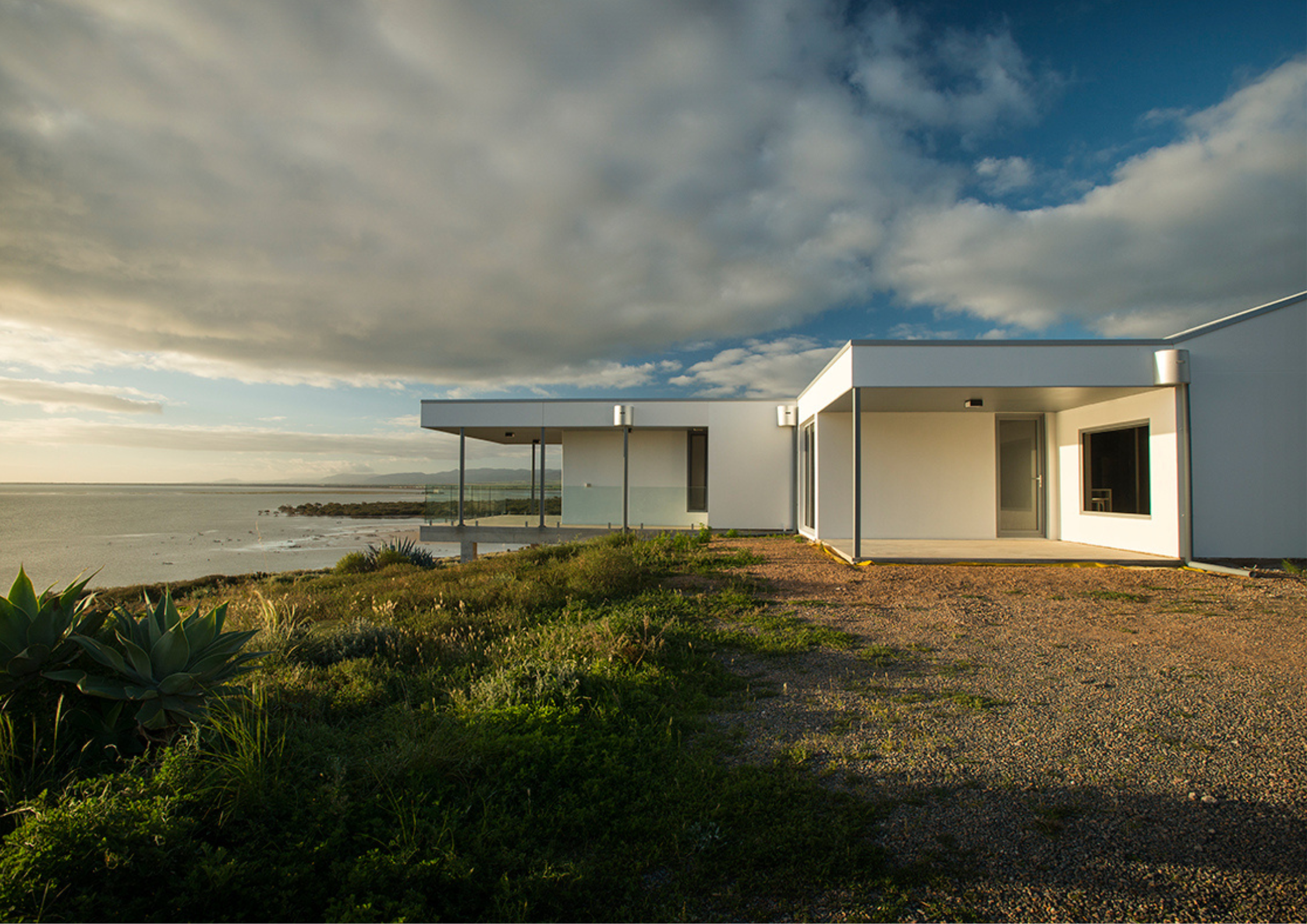 A low, modern coastal house overlooking the ocean with white walls and expansive glazing under a dramatic sky.