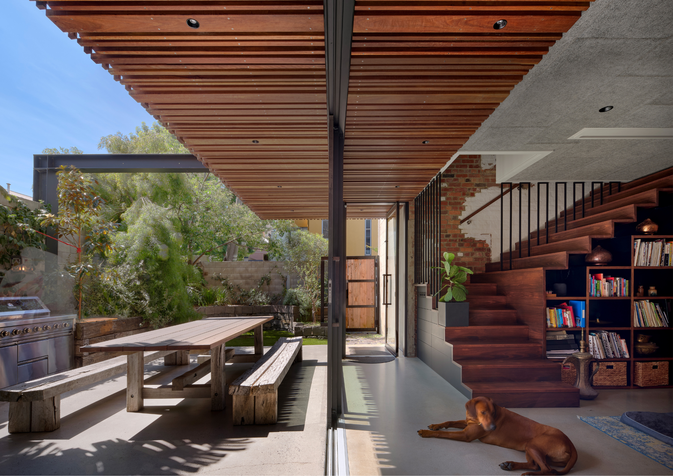 An indoor-outdoor space with a timber dining table, slatted ceiling, open glass wall, and a timber staircase with shelving.