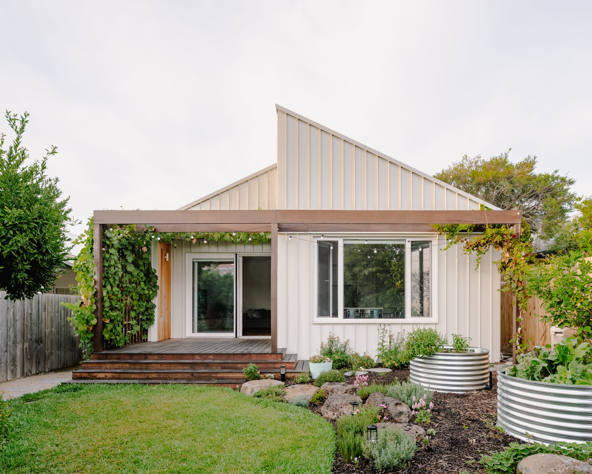 Light-toned gable-roofed home with a timber pergola, productive garden beds, and landscaped backyard.