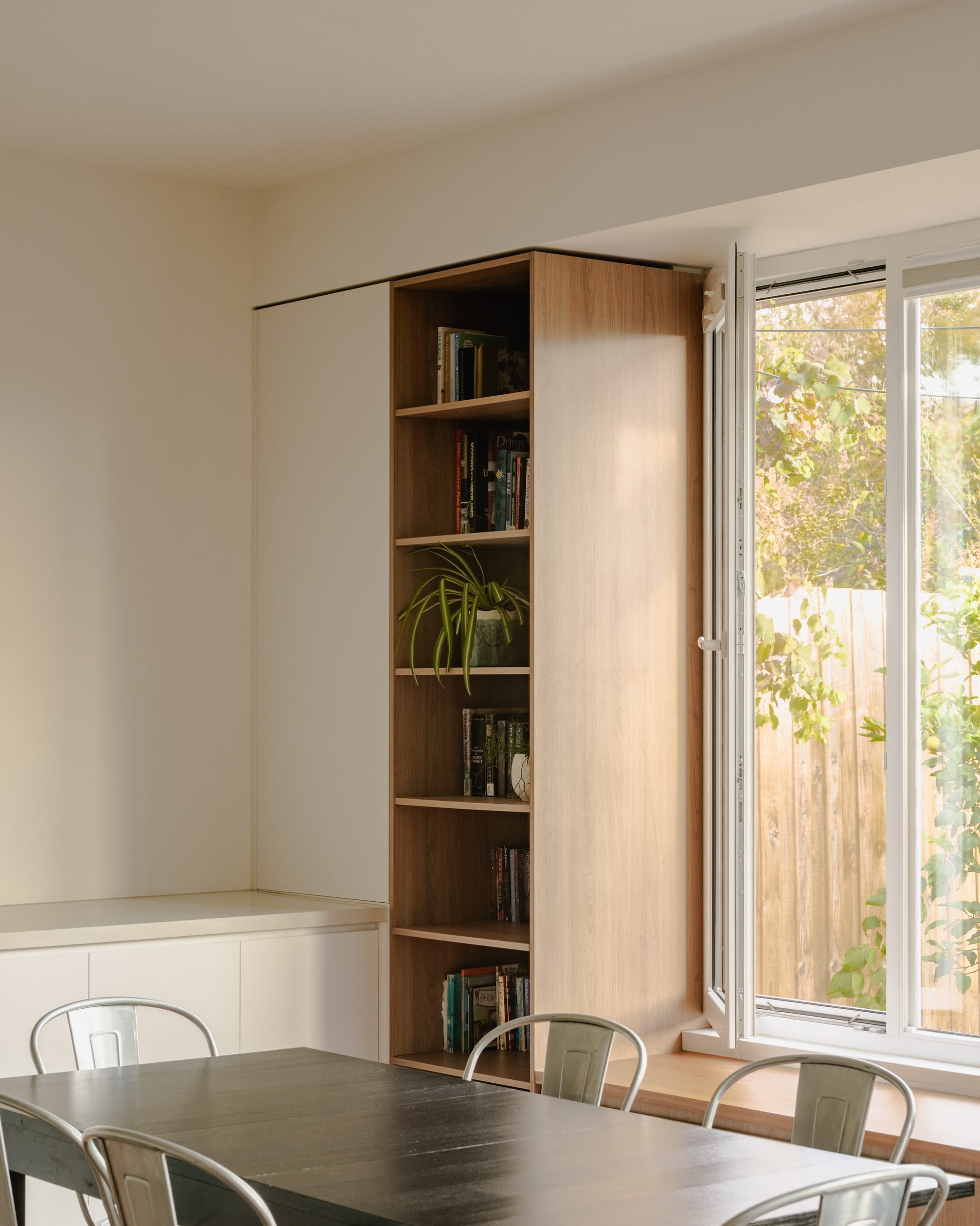 Minimal dining space with built-in timber shelving, white cabinetry, and an open window overlooking greenery.