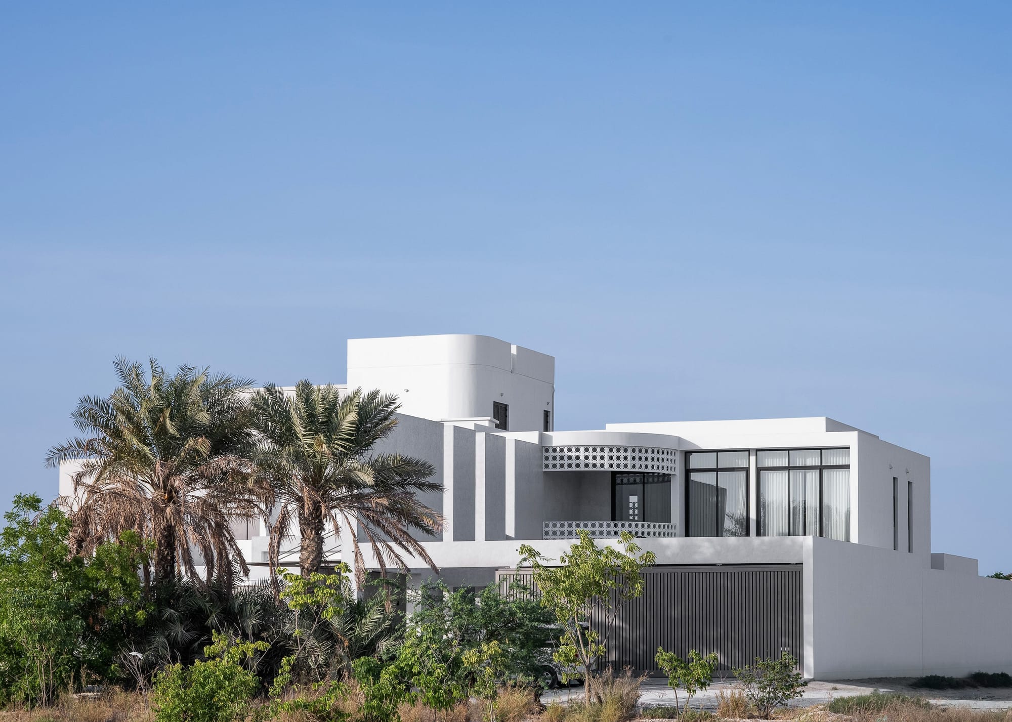 Contemporary white residence with geometric forms, screened balcony details and surrounding palm trees under a clear blue sky.
