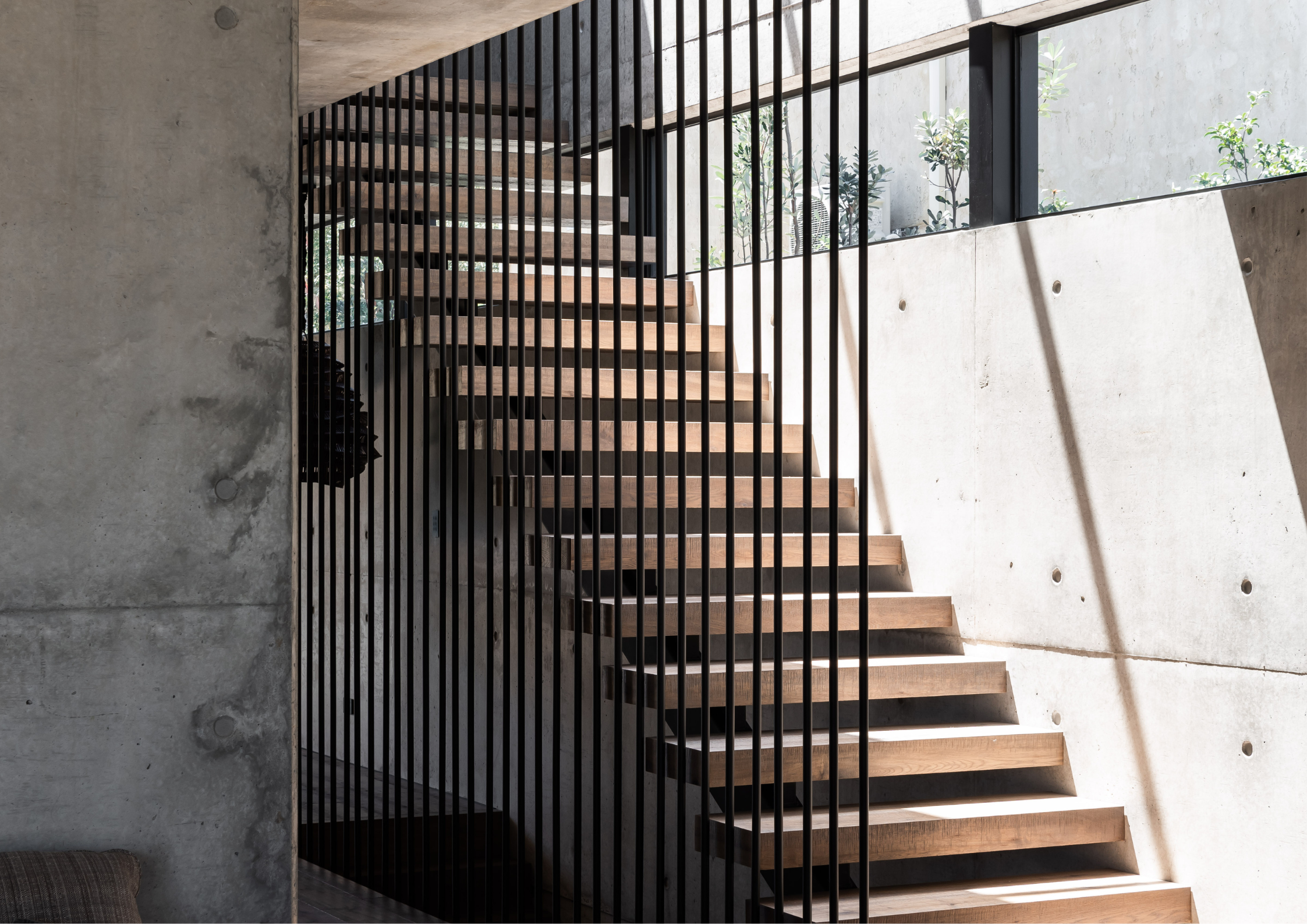 Sculptural timber staircase framed by black vertical balustrades and raw concrete walls, illuminated by natural light from above.