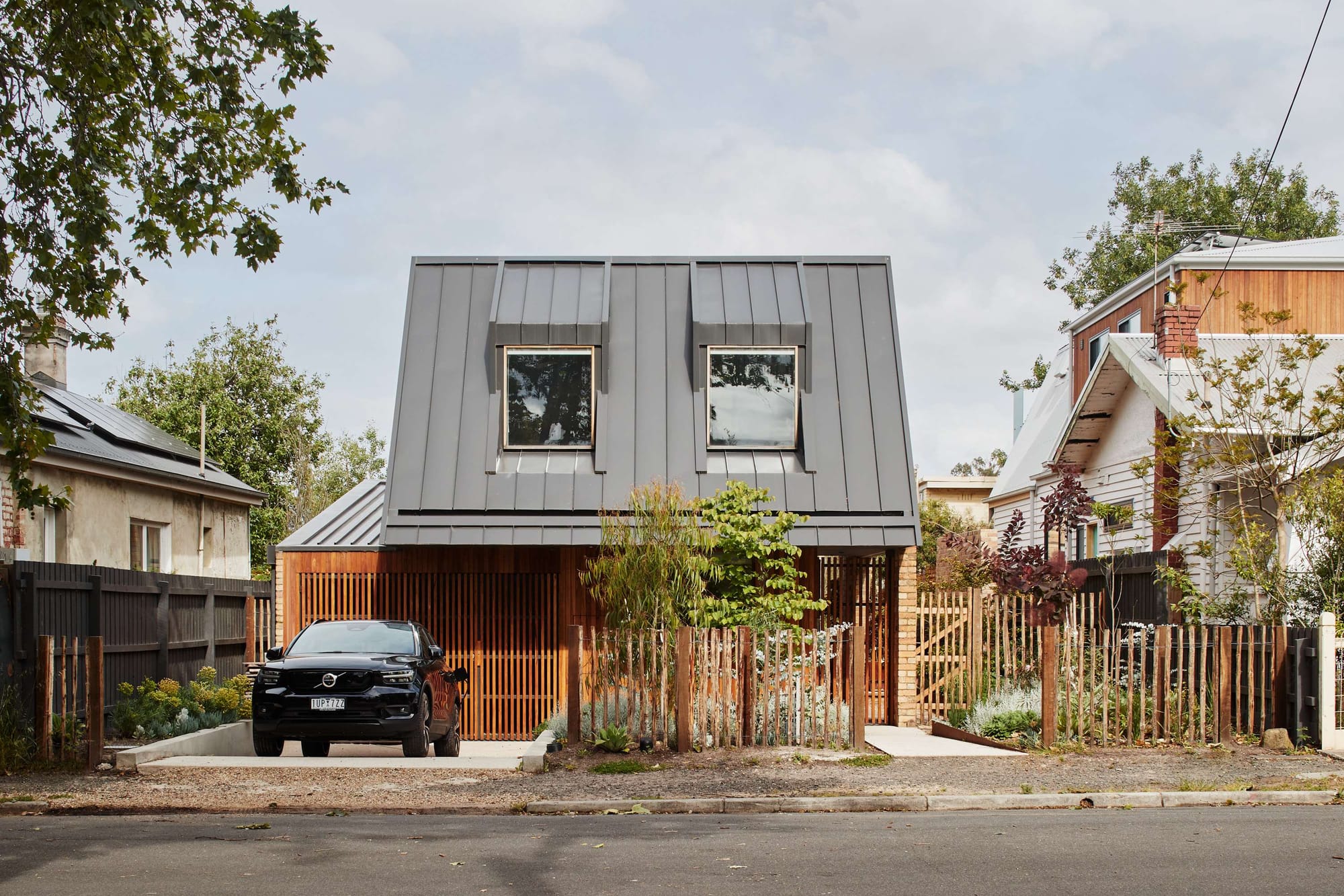 Contemporary compact home with dark standing-seam metal cladding, timber screening, and native landscaping set within a suburban streetscape.