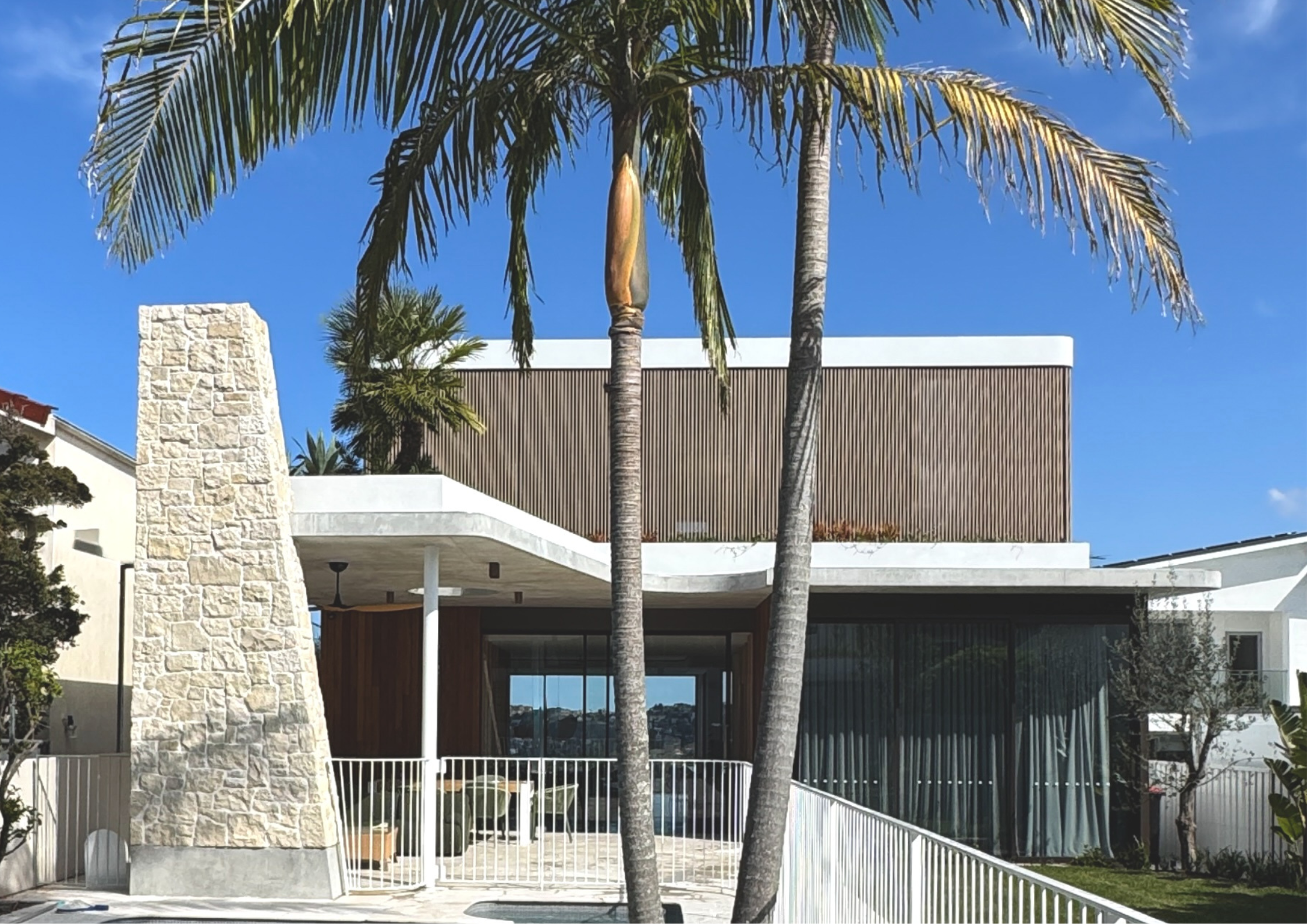 Modern coastal-inspired residence featuring curved concrete forms, timber screening, and tropical landscaping framed by tall palm trees.