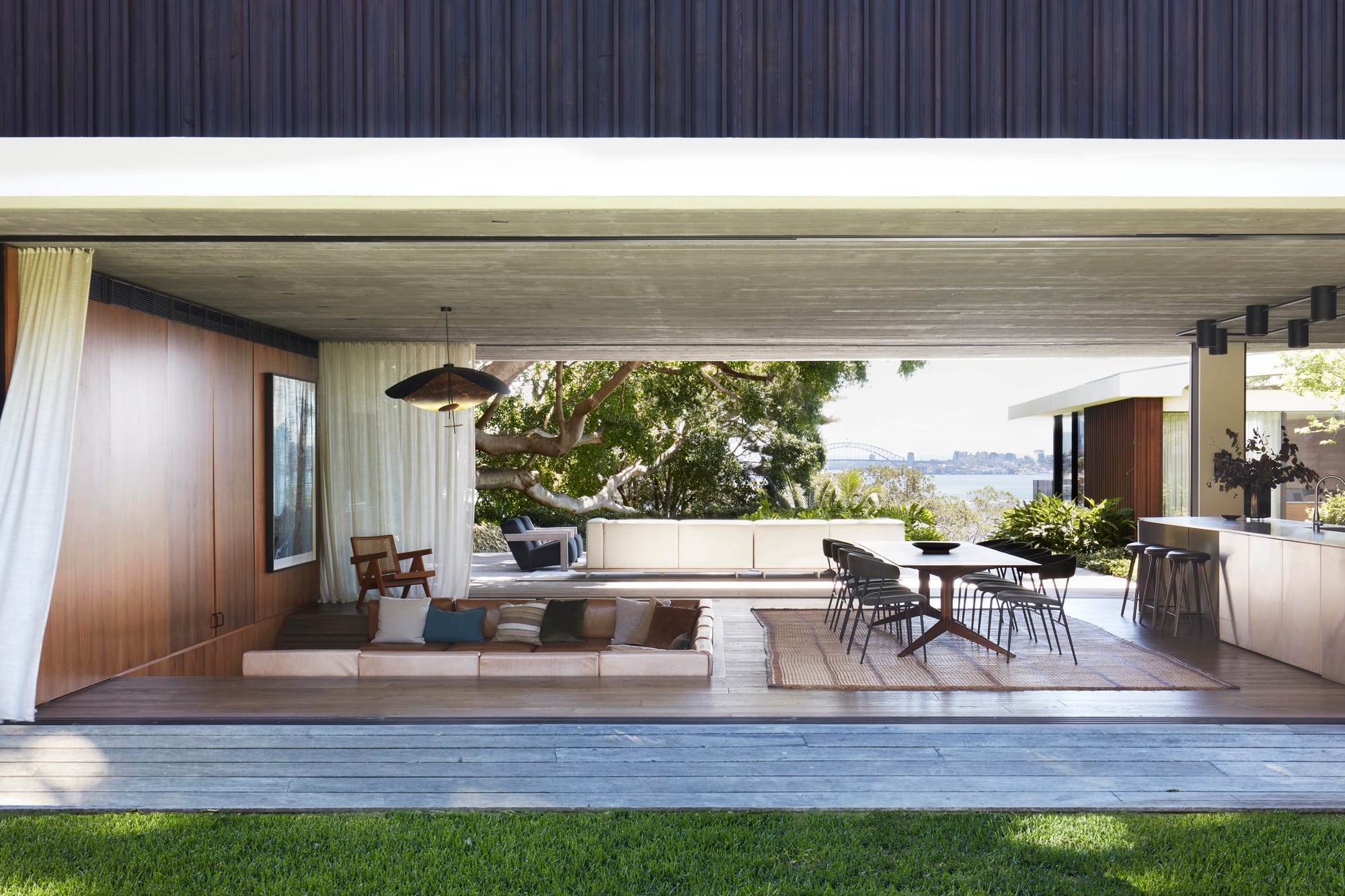 Open-plan indoor-outdoor living pavilion with timber finishes, sunken lounge, and expansive views toward Sydney Harbour through mature trees.