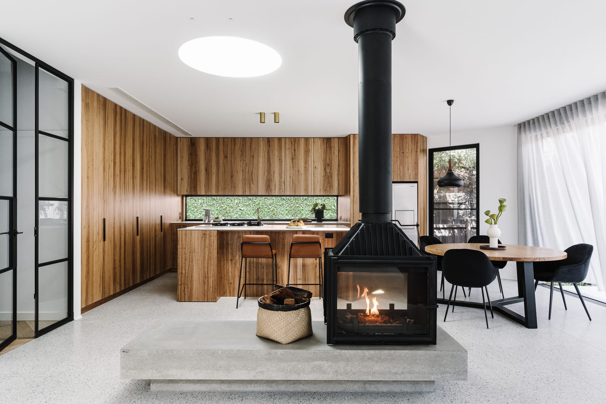 Minimal timber kitchen and dining space centred around a suspended black fireplace, with terrazzo flooring and soft natural daylight throughout.