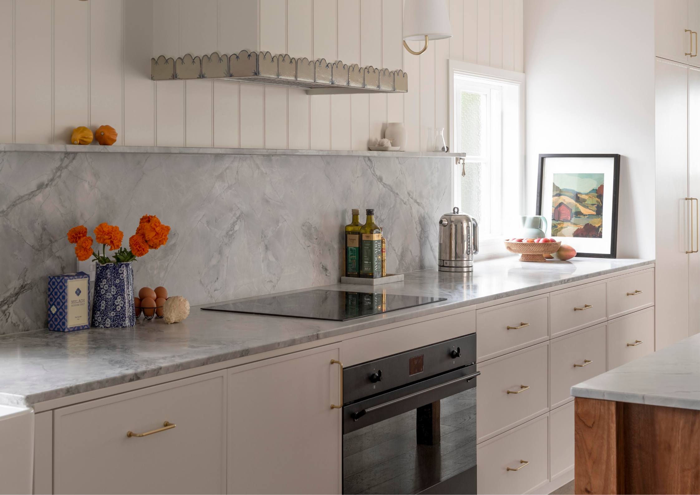 Bright, minimalist kitchen with pale cabinetry, grey marble benchtops and splashback, brass hardware and warm timber accents, styled with orange flowers, framed artwork and natural light from a side window.
