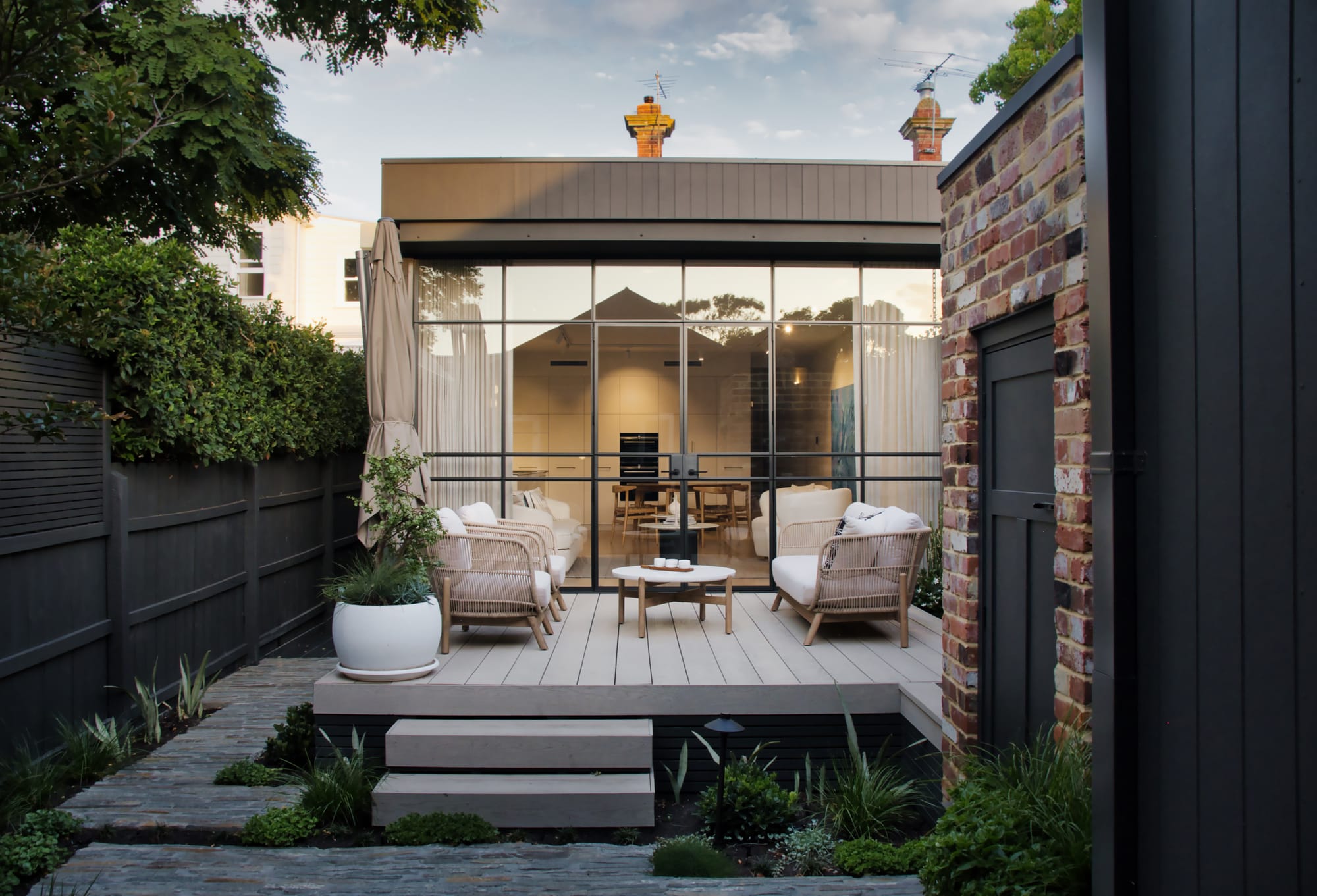 Outdoor entertaining deck with timber decking, woven armchairs and a round coffee table, framed by a steel and glass facade opening to a lit interior, with brick wall and garden planting at dusk.