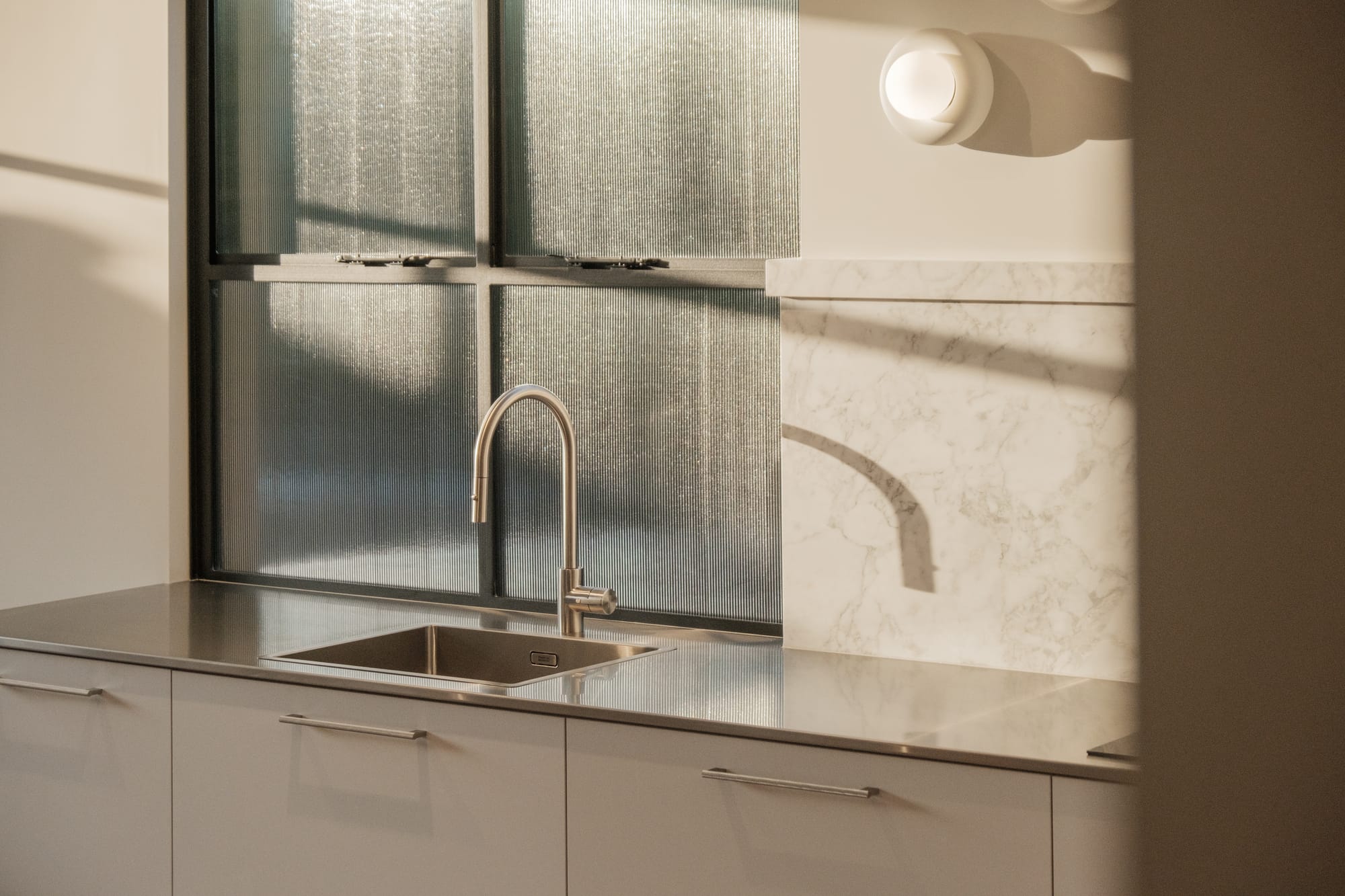 Close-up of a kitchen sink with a brushed steel tap set against a ribbed glass splashback, with marble benchtop and white cabinetry lit by warm afternoon light.