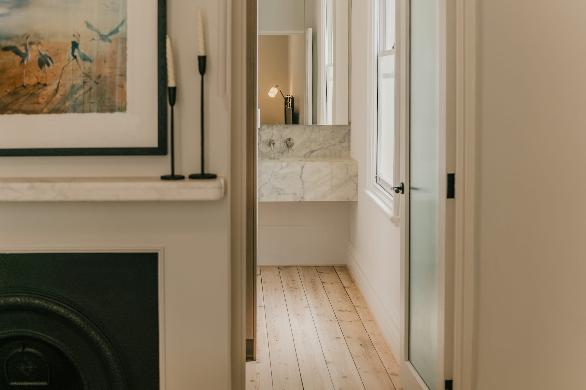 View through a doorway from a bedroom with a cast iron fireplace and artwork into an ensuite with a marble floating vanity, wall-mounted tapware, and light timber flooring.