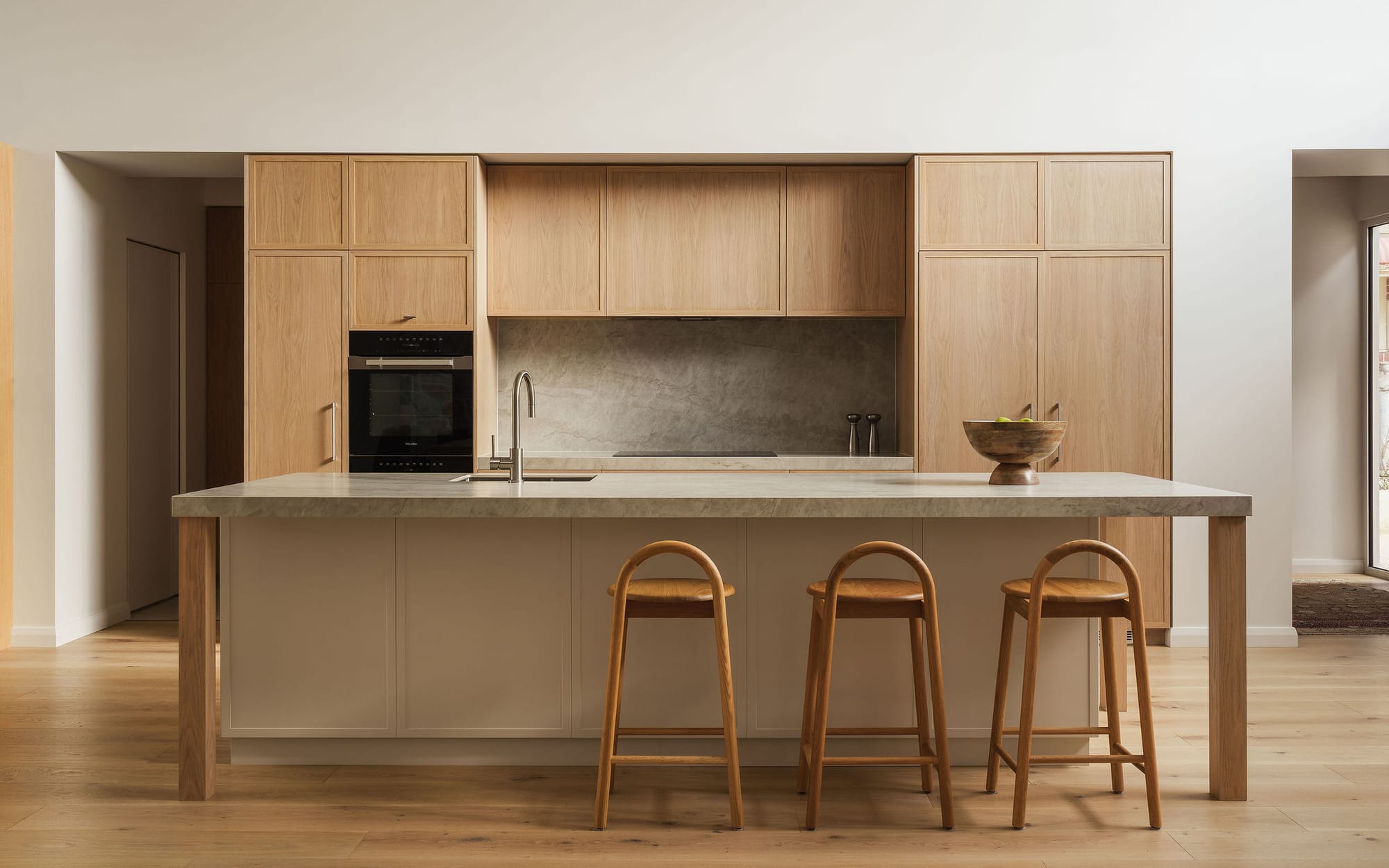  Bright open kitchen with full-height oak cabinetry, a stone island bench with timber legs and three oak stools, slate splashback, and light timber flooring in an open plan setting.