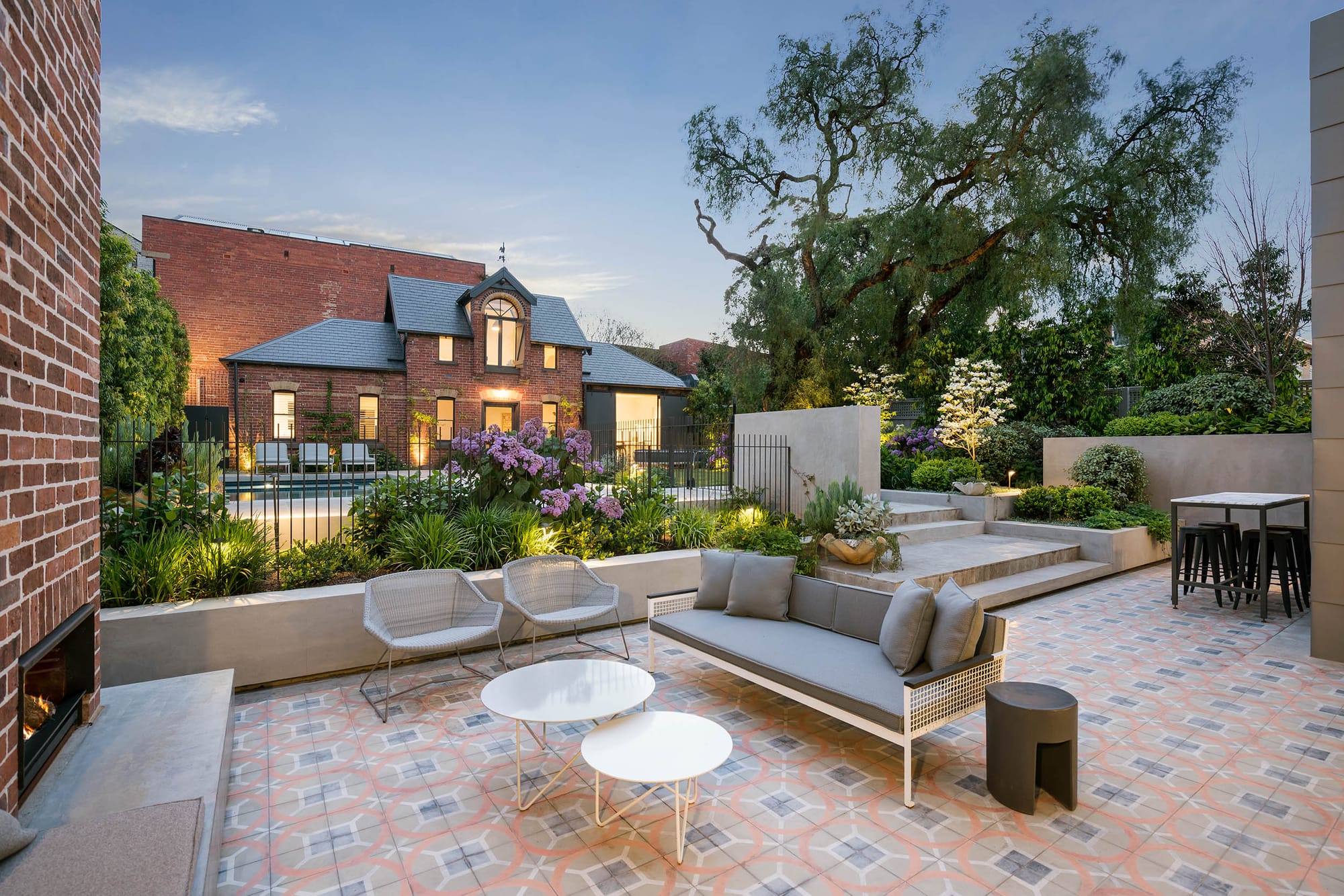 Outdoor entertaining terrace at the Armadale residence landscape by Myles Baldwin Design, with encaustic patterned tiles, lounge seating, a built-in fireplace and lush garden planting stepping up to the pool level beyond