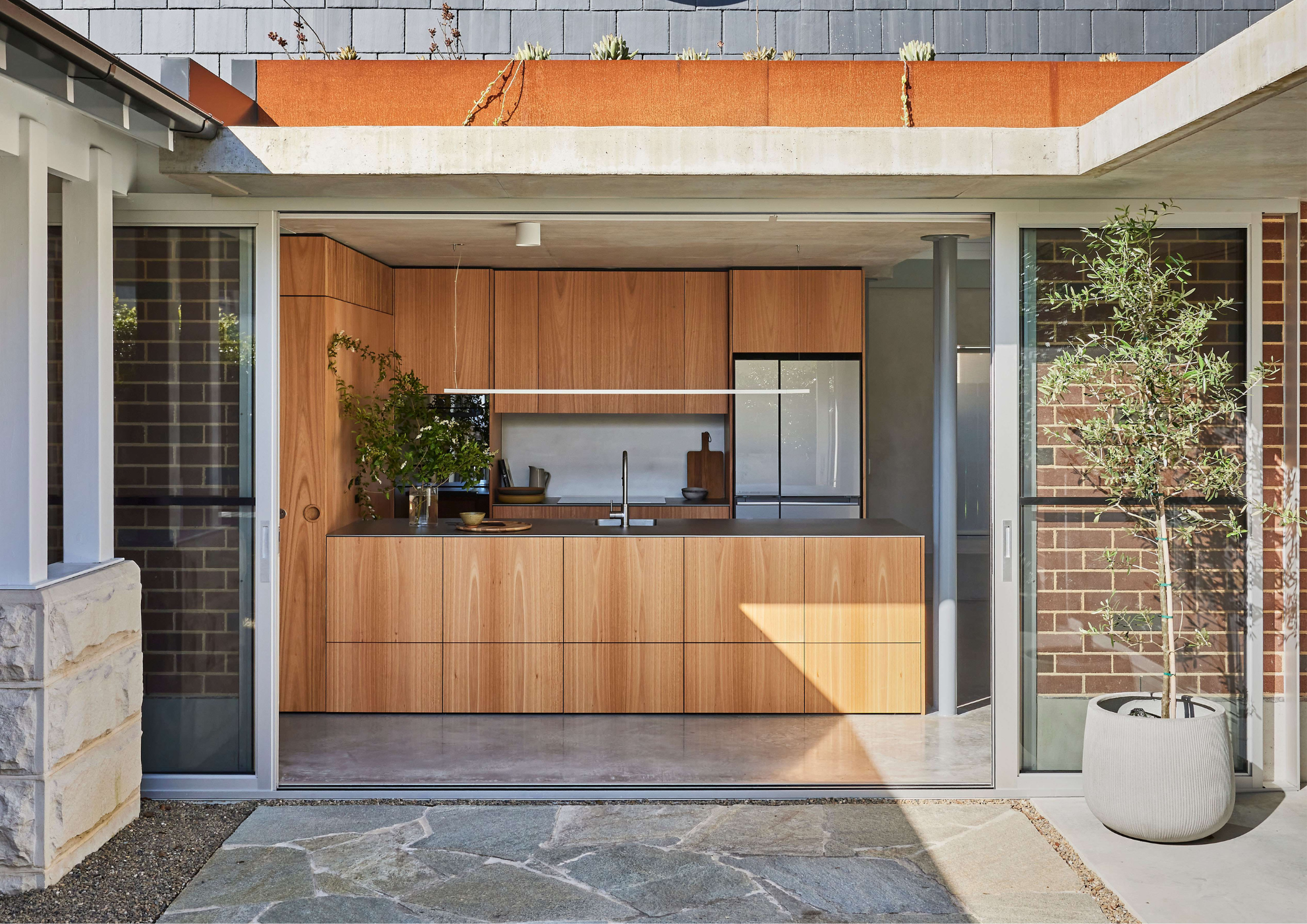 Timber kitchen opening to a courtyard in Courtyard House by Atelier M, with sliding glass doors, stone paving, and a potted olive tree connecting interior and exterior.