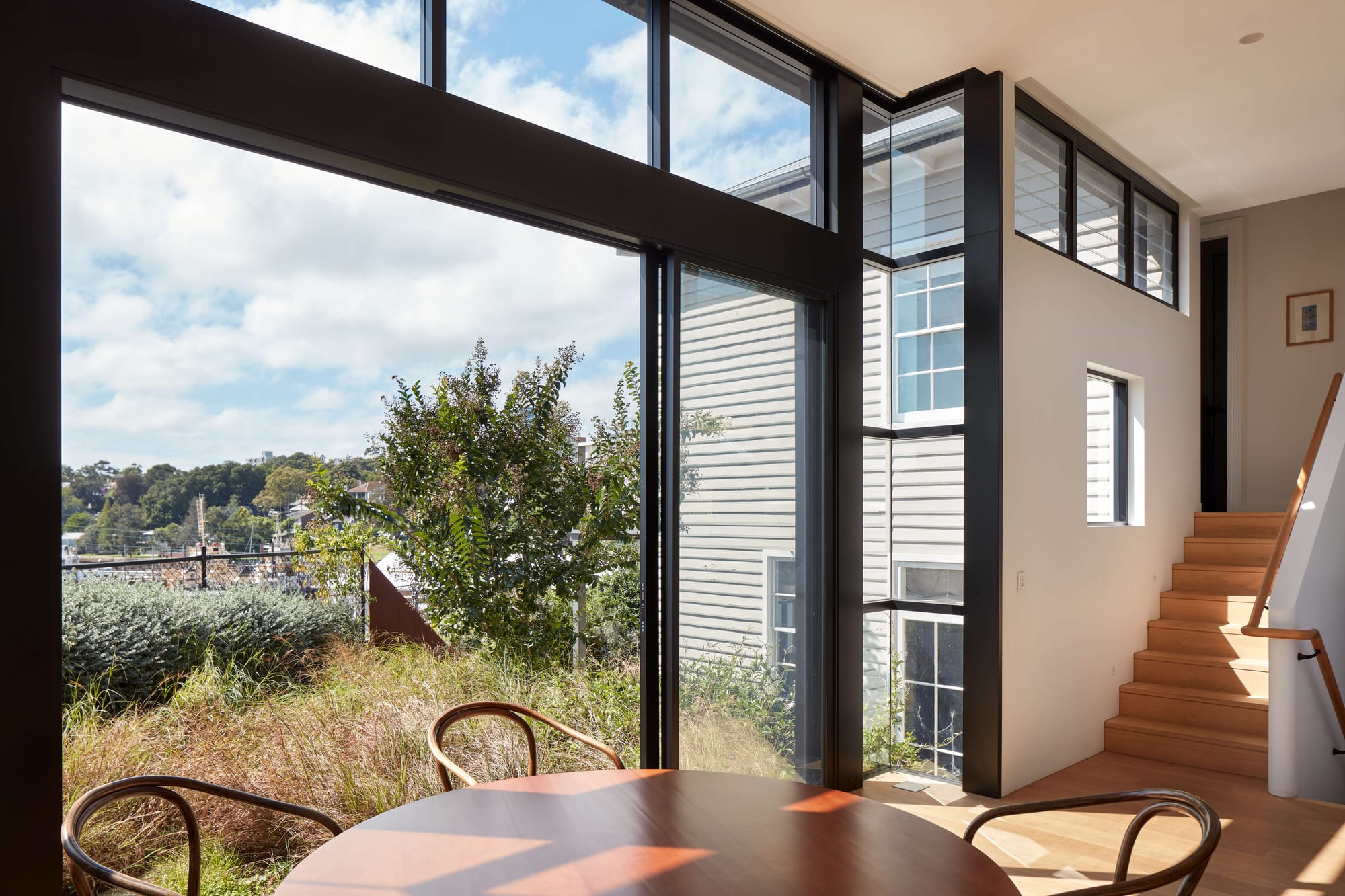 Floor-to-ceiling black-framed glazing in the Balmain House by Corso Architecture, framing harbour and garden views from the dining area with a timber staircase beyond.