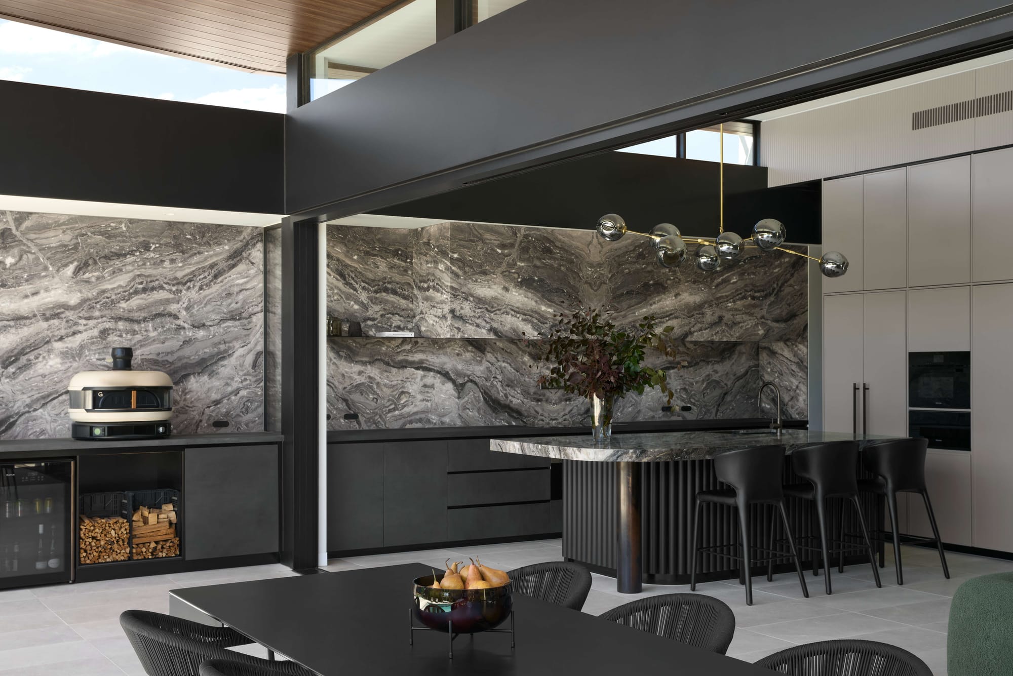 A dramatic outdoor kitchen with floor to ceiling stone slab splashback and marble island bench opens to an alfresco dining area beneath a dark steel pergola, Balwyn Residence by Karavasil Architects