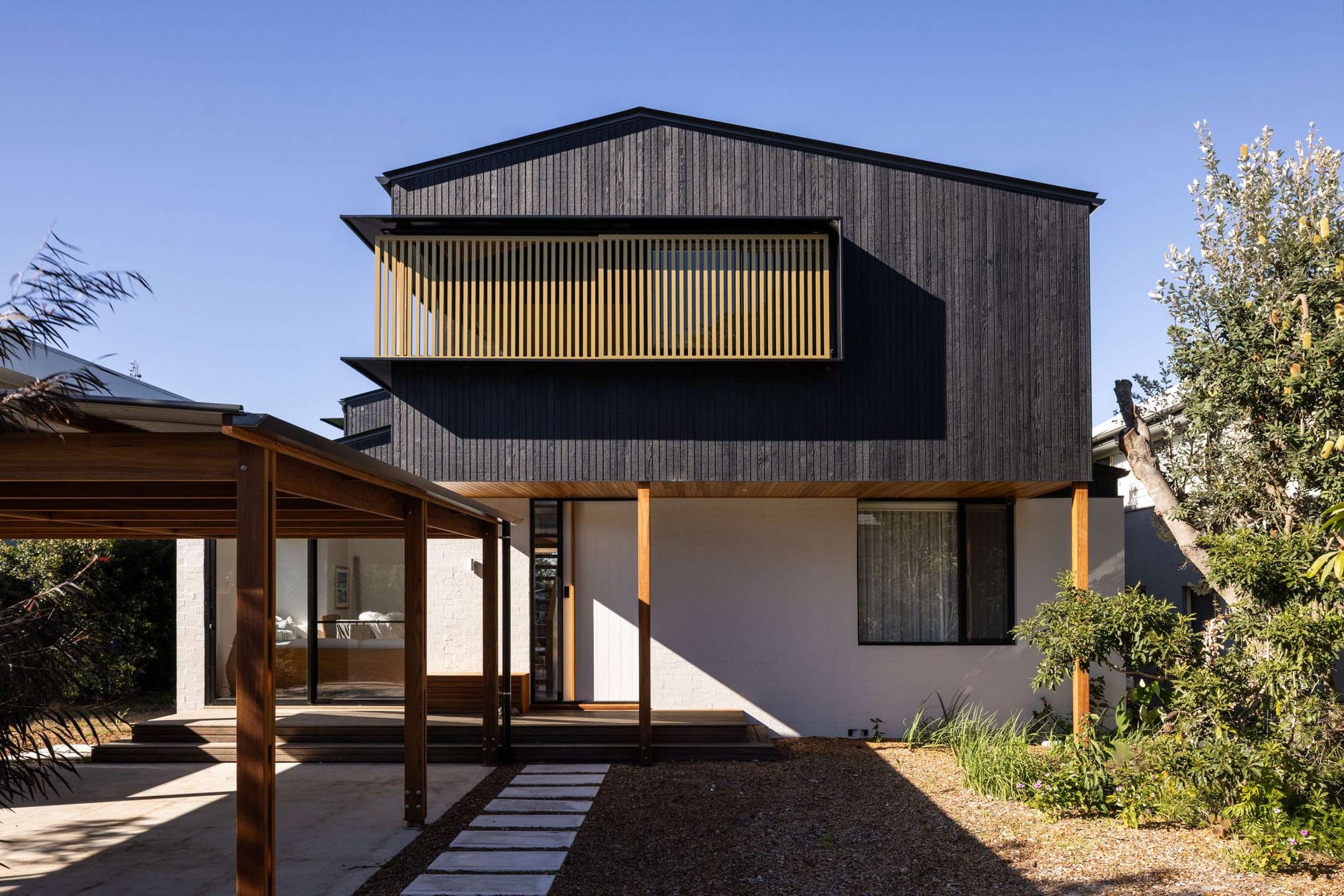 A timber pergola structure connects a landscaped garden to the living areas of a two storey home clad in dark charred timber, Bawley Point House by Watershed Architects