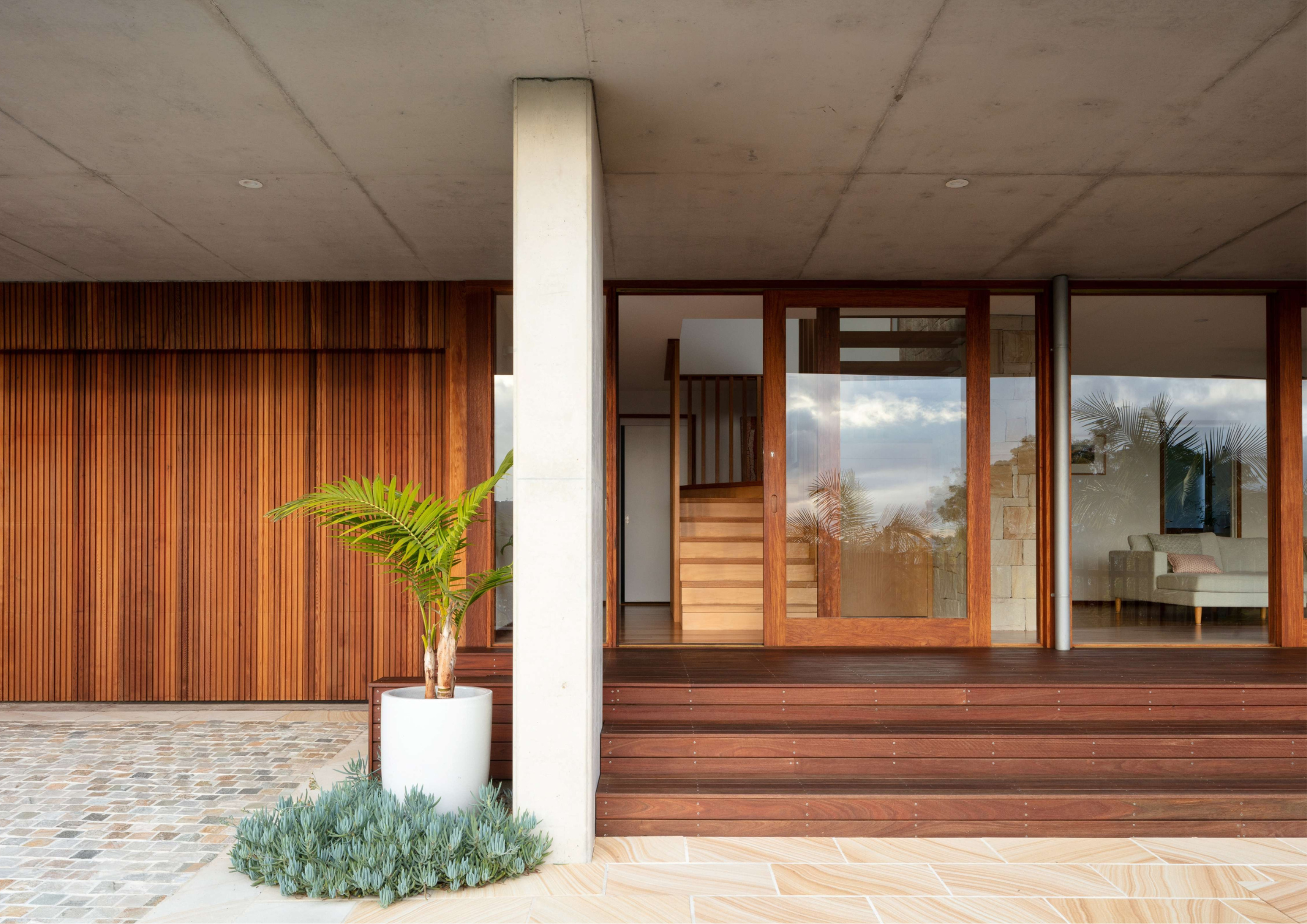 Elevated hardwood deck with exposed concrete soffit, timber batten wall cladding, large glazed doors, and a potted palm in the foreground.
