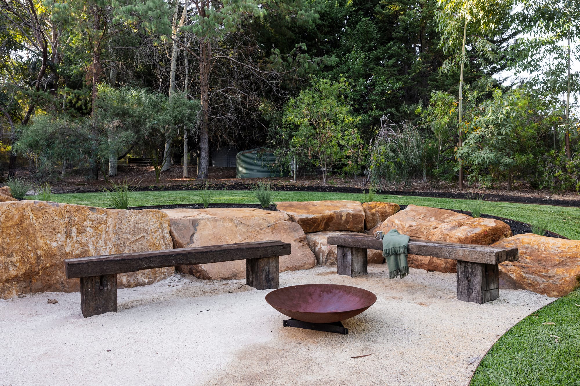 Outdoor fire pit area with a corten steel bowl, rustic timber benches and large natural sandstone boulders set against a lawn and native tree backdrop.