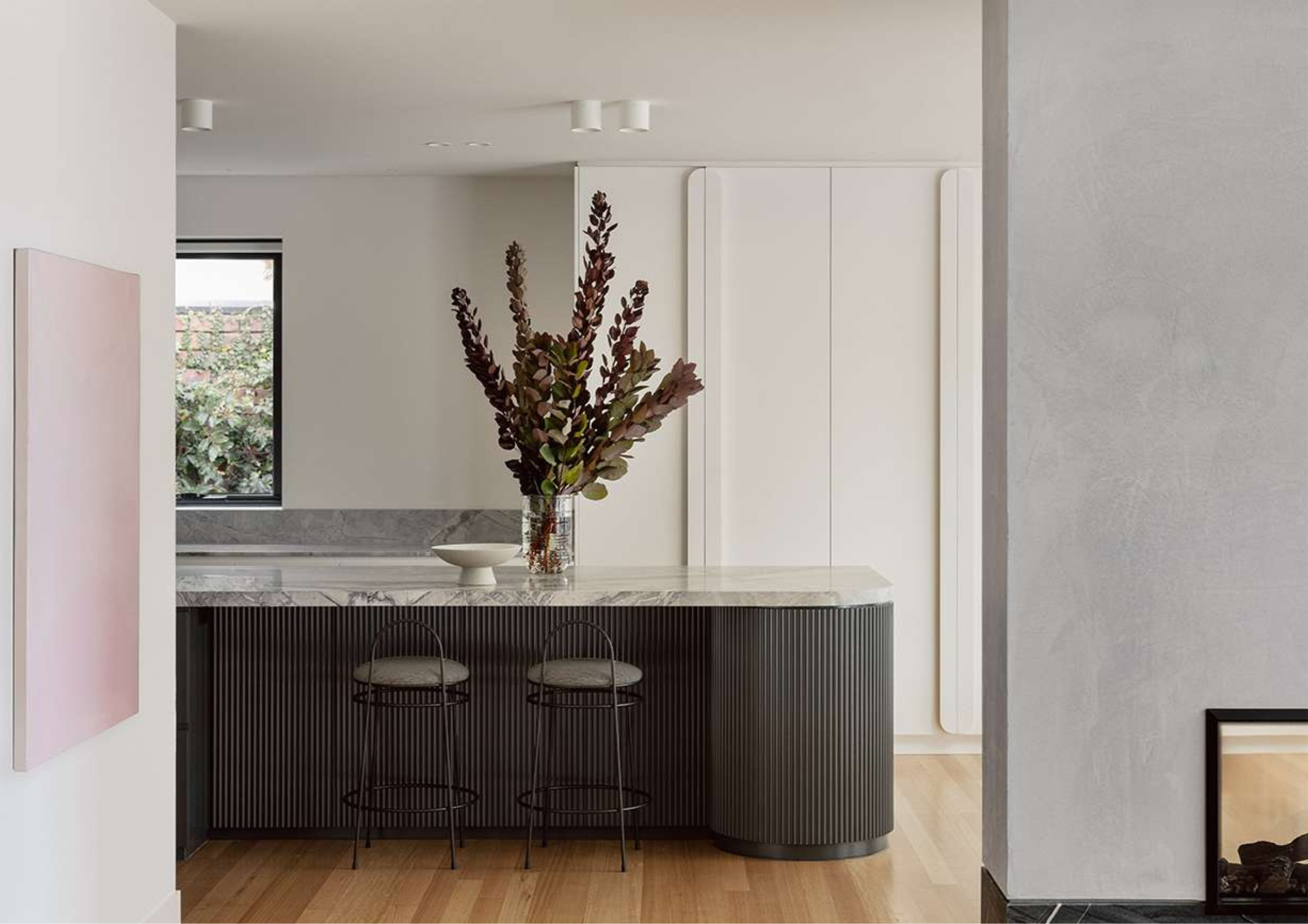 Marble kitchen island with fluted dark cabinetry and bar stools in the Caulfield Project by SMD Projects, with a feature wall panel and garden views through a black-framed window.