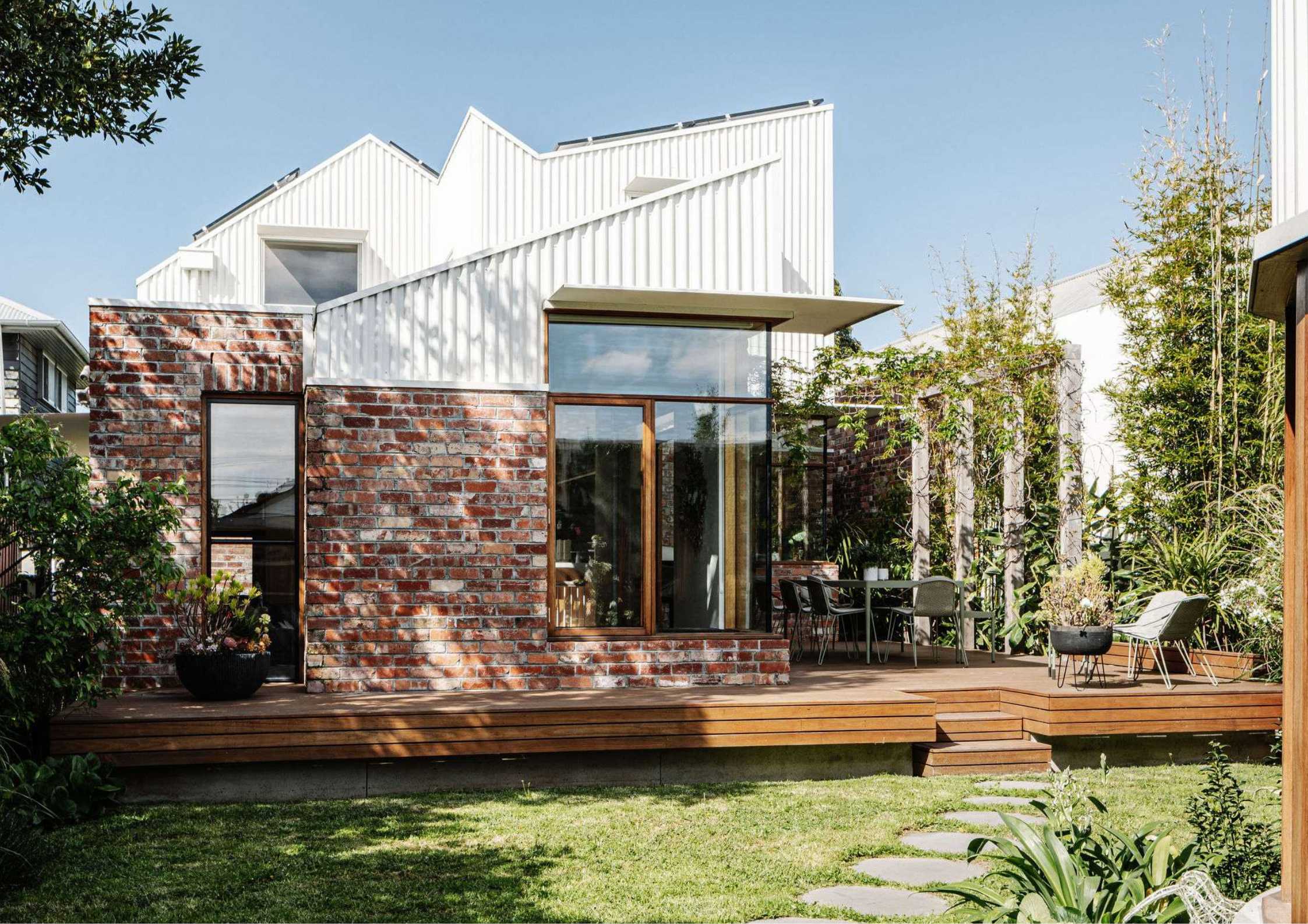 Hardwood timber deck extending from a recycled brick home with floor-to-ceiling glazing, outdoor dining setting, and lush garden landscaping.