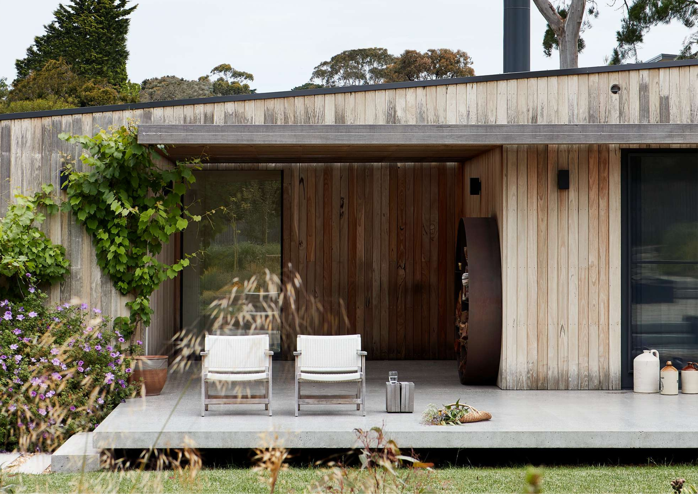 Concrete alfresco terrace with timber-clad walls, two outdoor lounge chairs, and a climbing vine beside sliding glass doors opening to a garden setting.