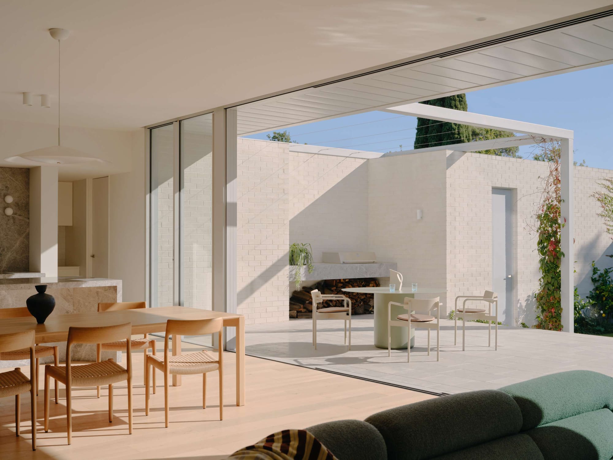 Stacking glass doors dissolve the boundary between a timber floored dining room and a white brick courtyard with outdoor seating and a fireplace, Coppin House by Tom Robertson Architects