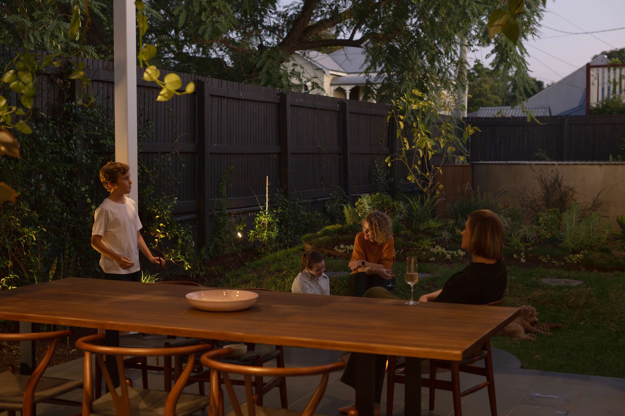 Family gathered at outdoor timber dining table at dusk surrounded by lush garden, Cornerstone by Loupe Architecture