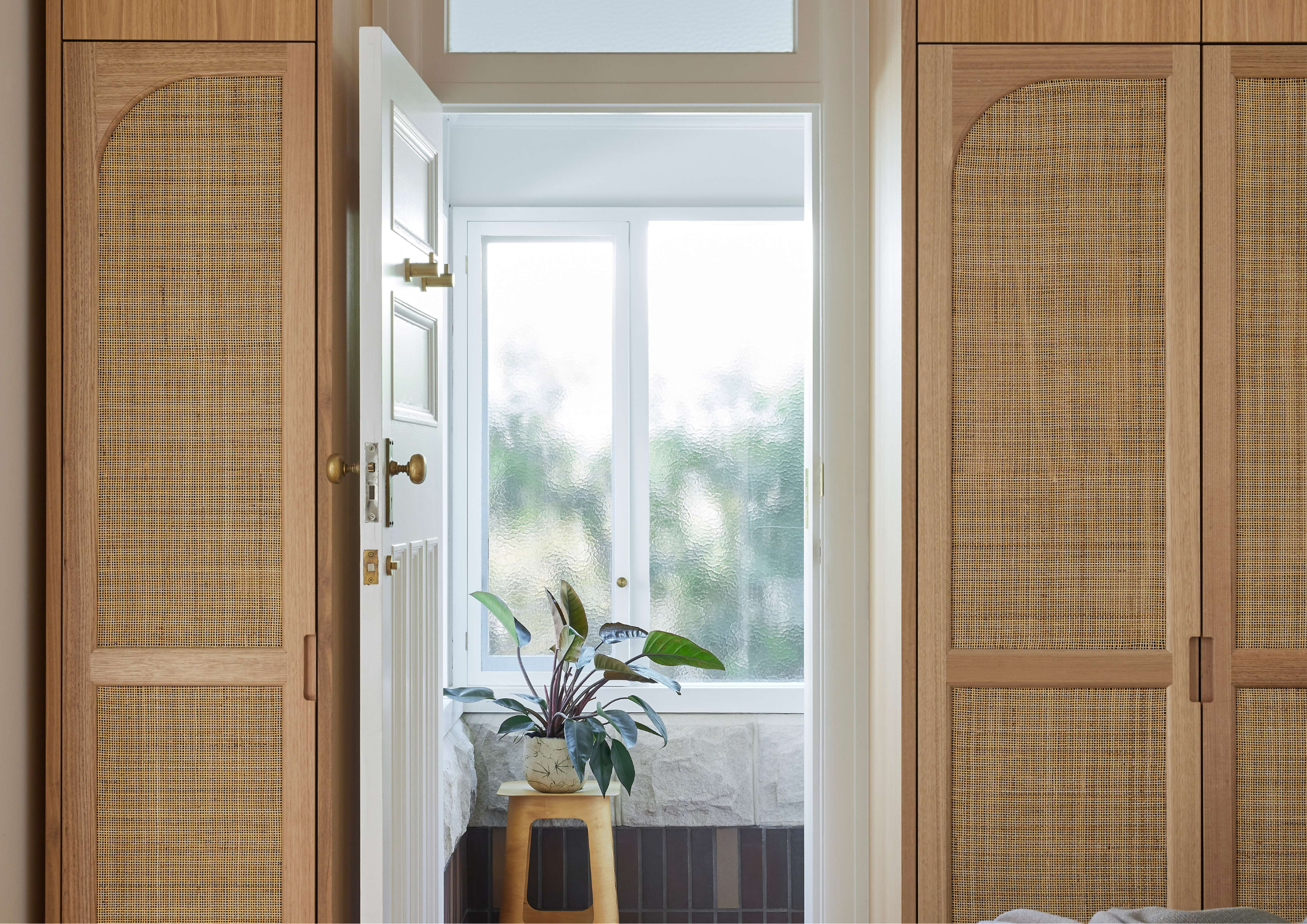 Hallway view through arched oak and cane wardrobes toward a bright bathroom with textured glass window and indoor plant in Courtyard House by Atelier M.