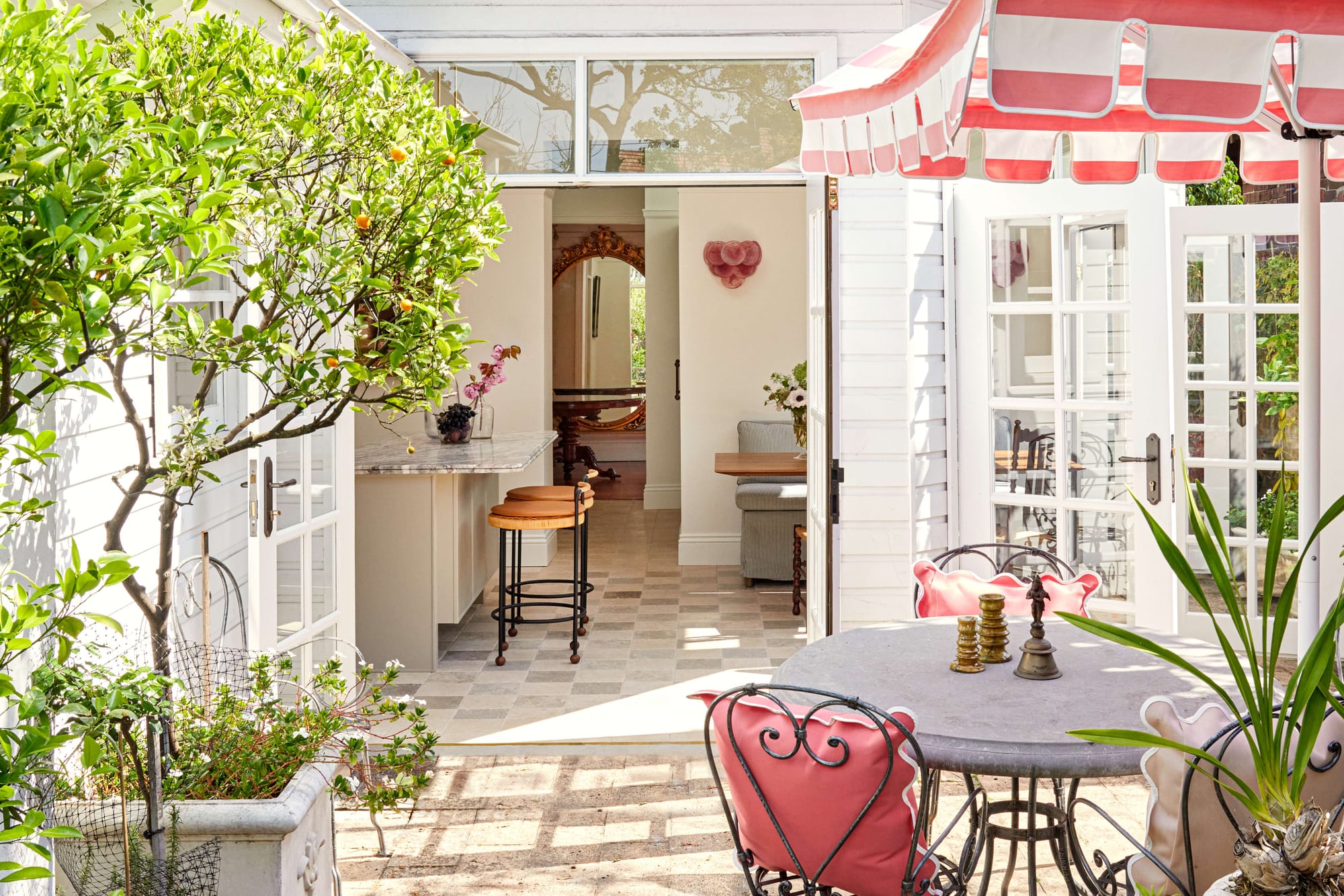 French doors open from a bright kitchen onto a courtyard dining area shaded by a striped umbrella and surrounded by citrus trees, Drummoyne Terrace by Studio PULP