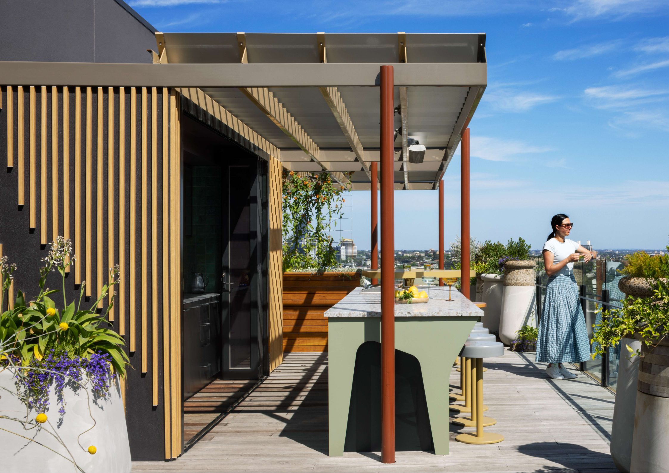 Timber-clad beach house with sliding glass doors opening to a covered alfresco with dining table, lounge seating, and built-in barbecue, overlooking a lawn and garden.