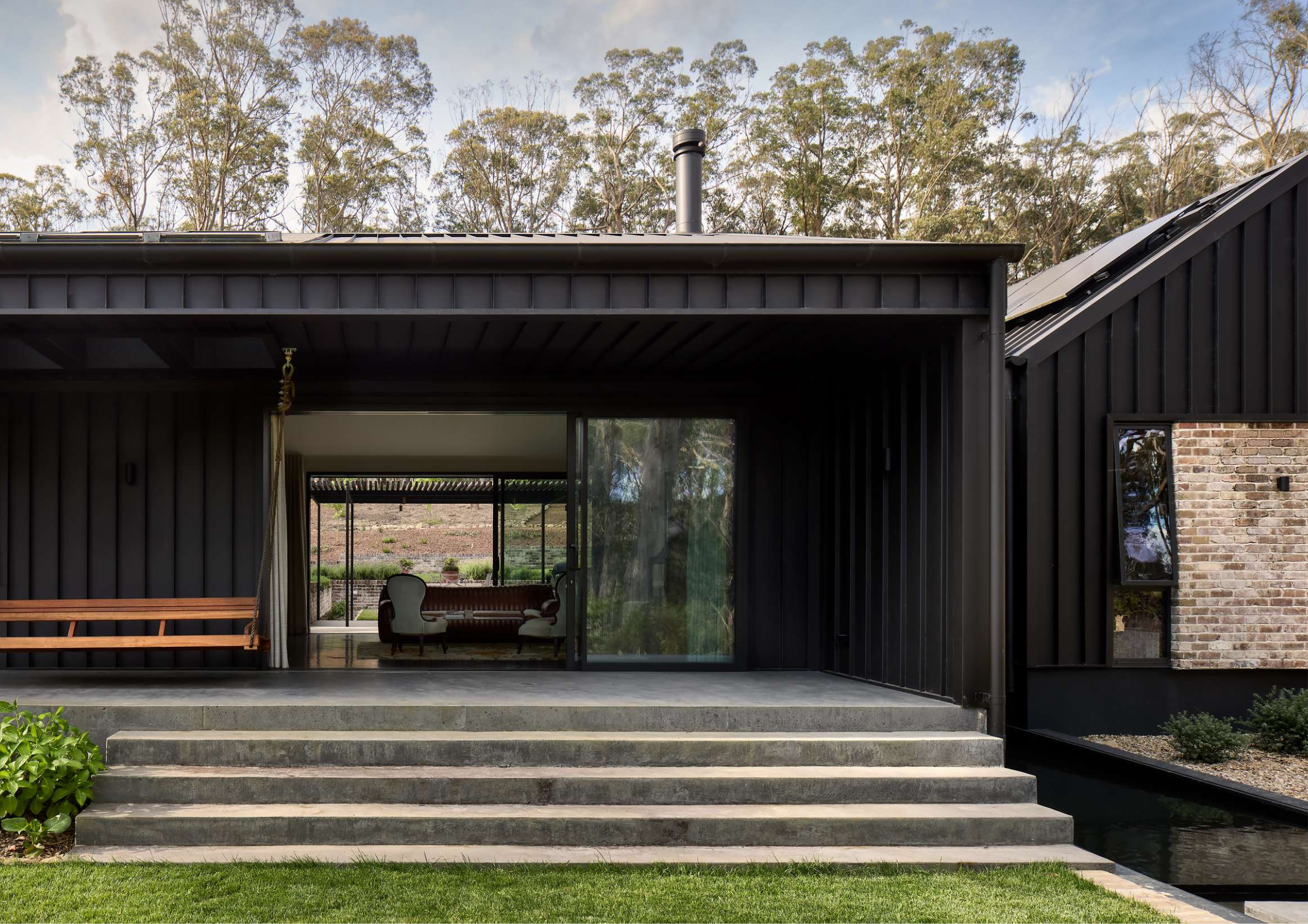 Dark steel-clad home with a covered alfresco, concrete steps leading to a sliding glass door, and a timber bench seat beside a lush garden.
