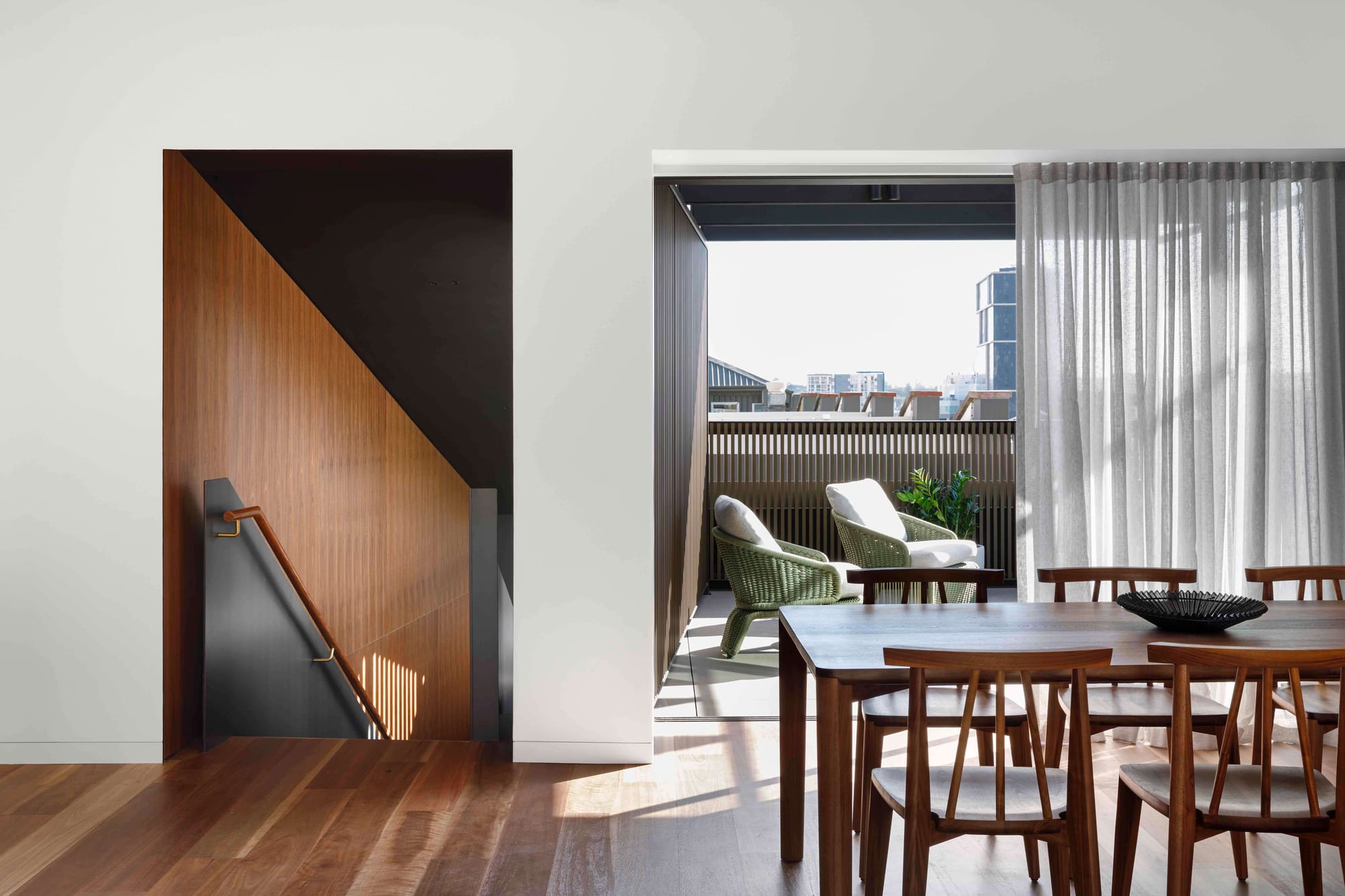 A dining area with a dark timber table and matching chairs opening through full-width glazed doors to a covered terrace with wicker lounge chairs, alongside a timber-lined stairwell with a brass handrail and warm afternoon light crossing the hardwood floor.