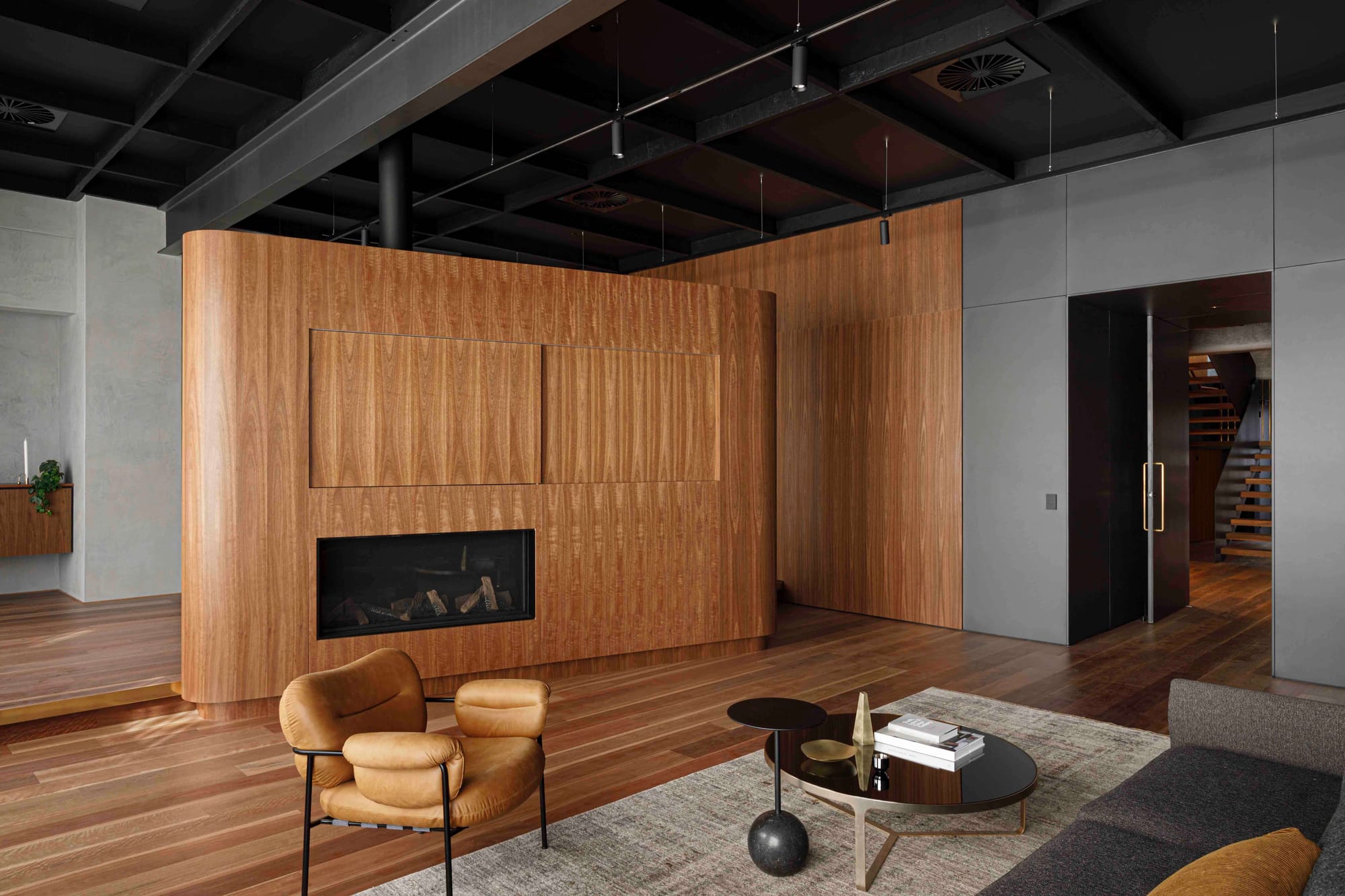A dramatic open-plan living space featuring a curved timber wall with integrated linear fireplace, exposed black structural ceiling with pendant track lighting, concrete-finish walls, a tan leather armchair, and paired circular black coffee tables on a textured rug.