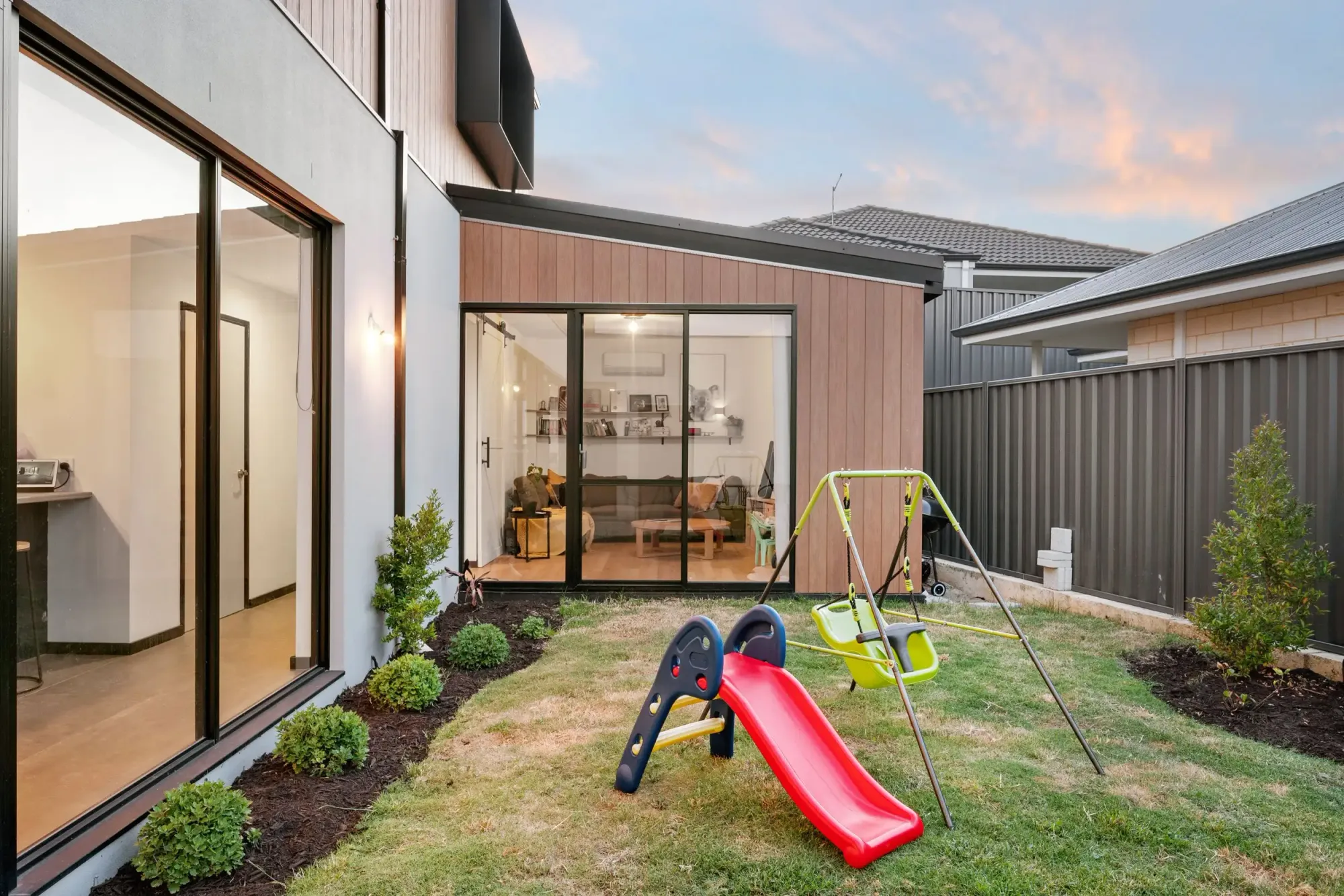 Children playing in backyard with swing set and slide, timber-clad addition and sliding glass doors, Koto House by Cynosure Architecture